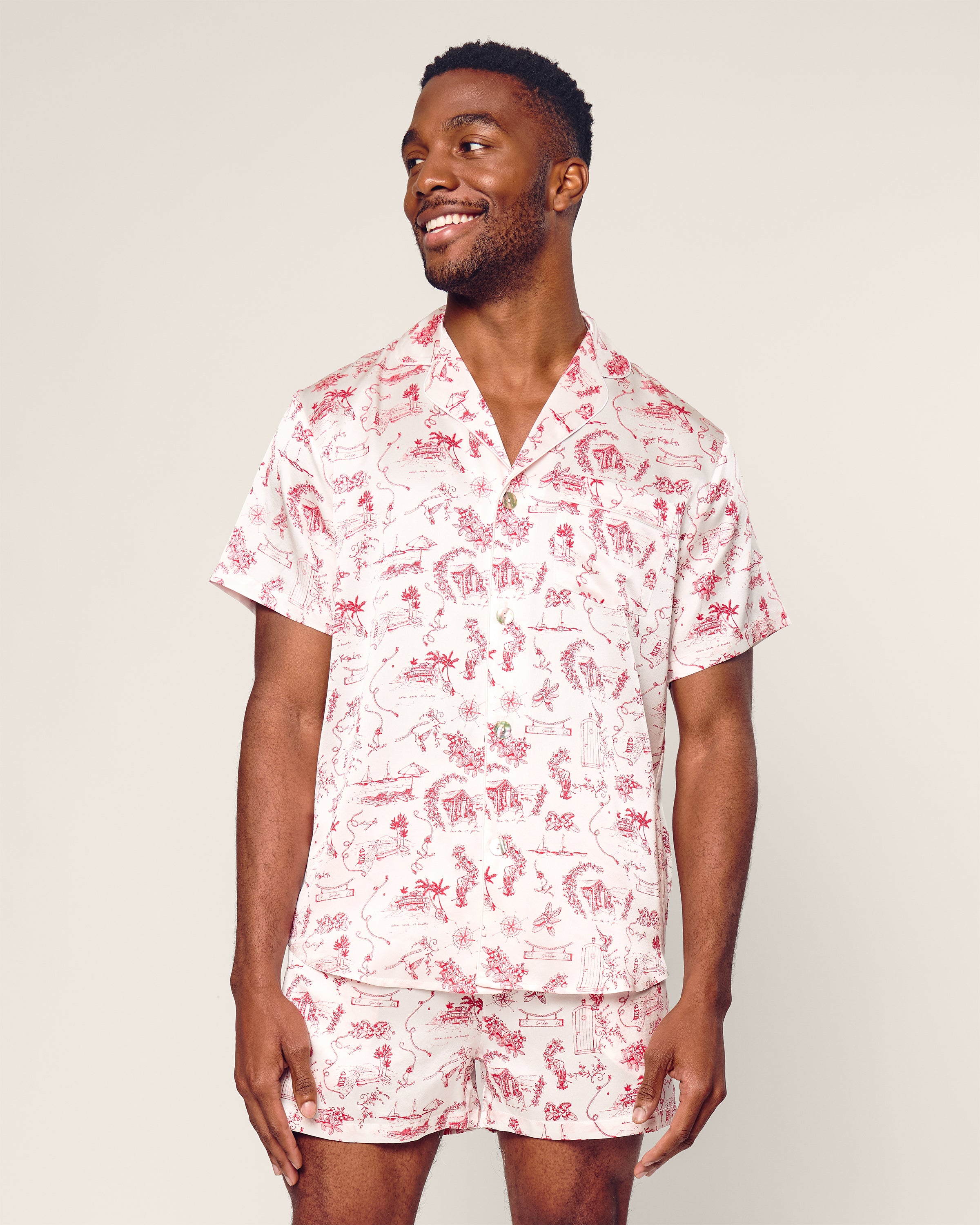 A man models the Petite Plume x Eden Rock Reverie Mens Silk Short Set, featuring a red and white print. He stands against a simple backdrop, smiling and gazing to the side, capturing relaxed St Barths vibes.