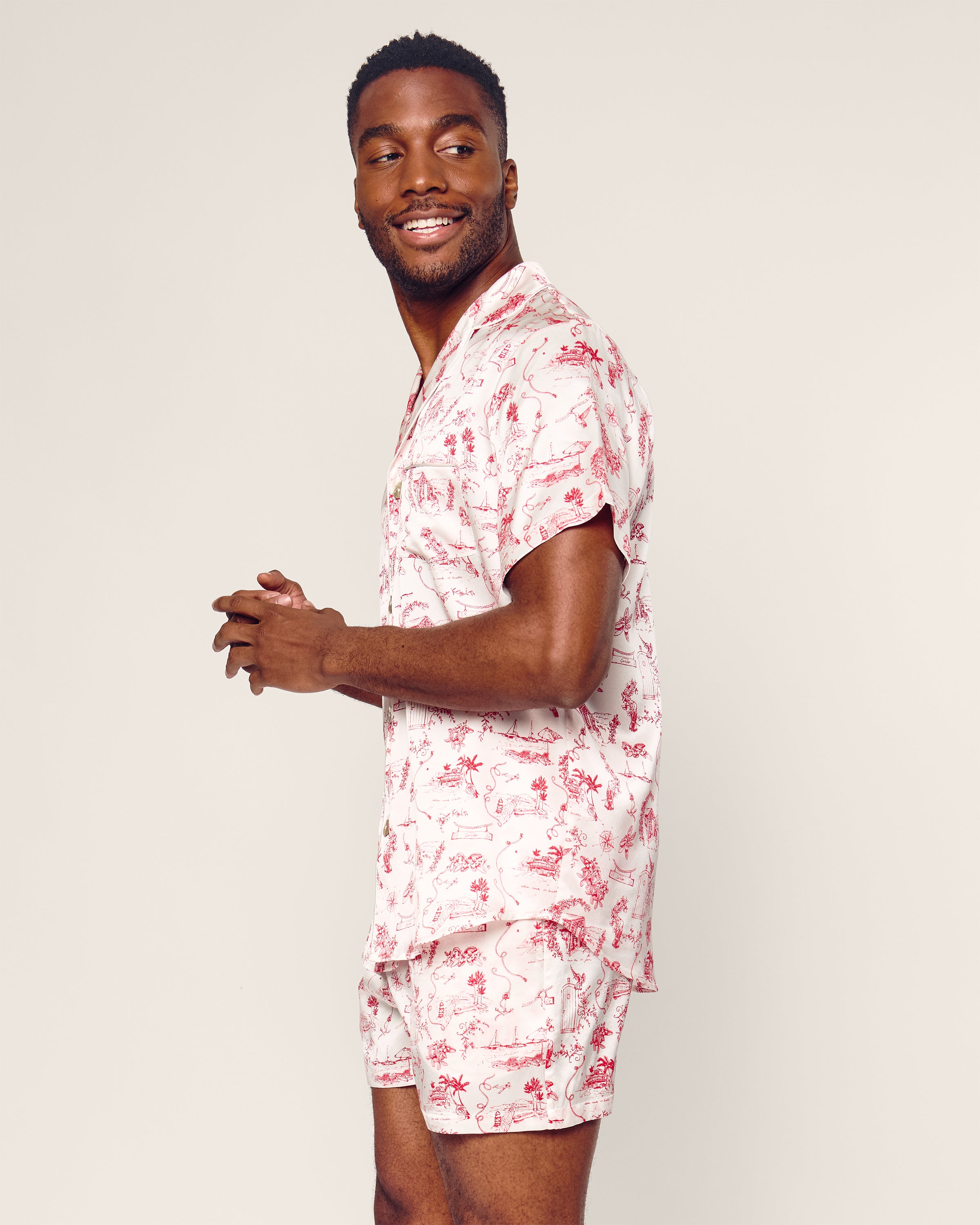 A person smiles in a Petite Plume x Eden Rock Reverie Mens Silk Short Set, showcasing a red and white floral pattern. This chic summer ensemble from Petite Plume evokes the relaxed spirit of St Barths, standing out against a plain light beige background.