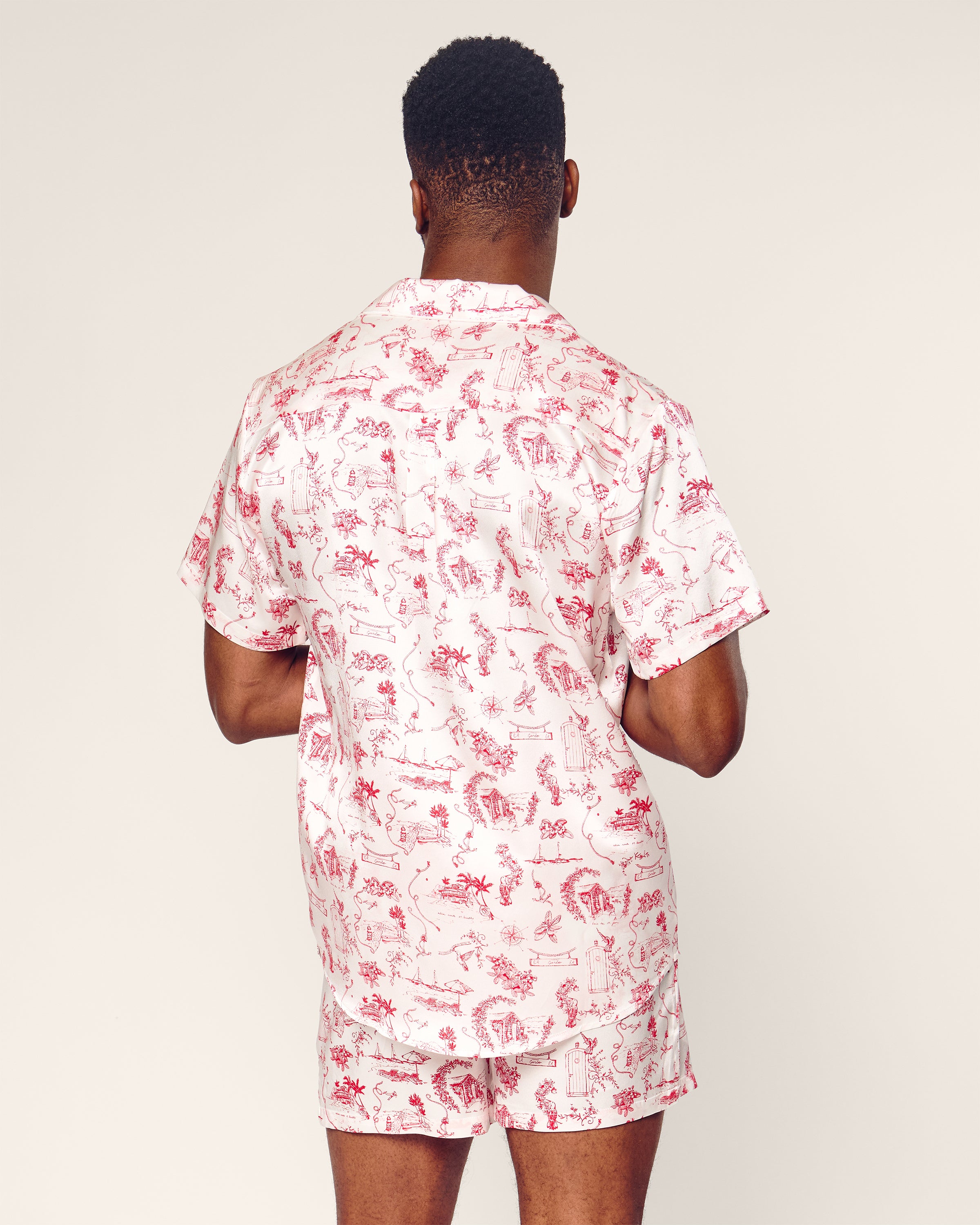 A person wearing the Petite Plume x Eden Rock Reverie Mens Silk Short Set, featuring a red and white intricate pattern, stands with back to the camera. The luxury sleepwear showcases the elegance of mulberry silk against a plain light beige background.