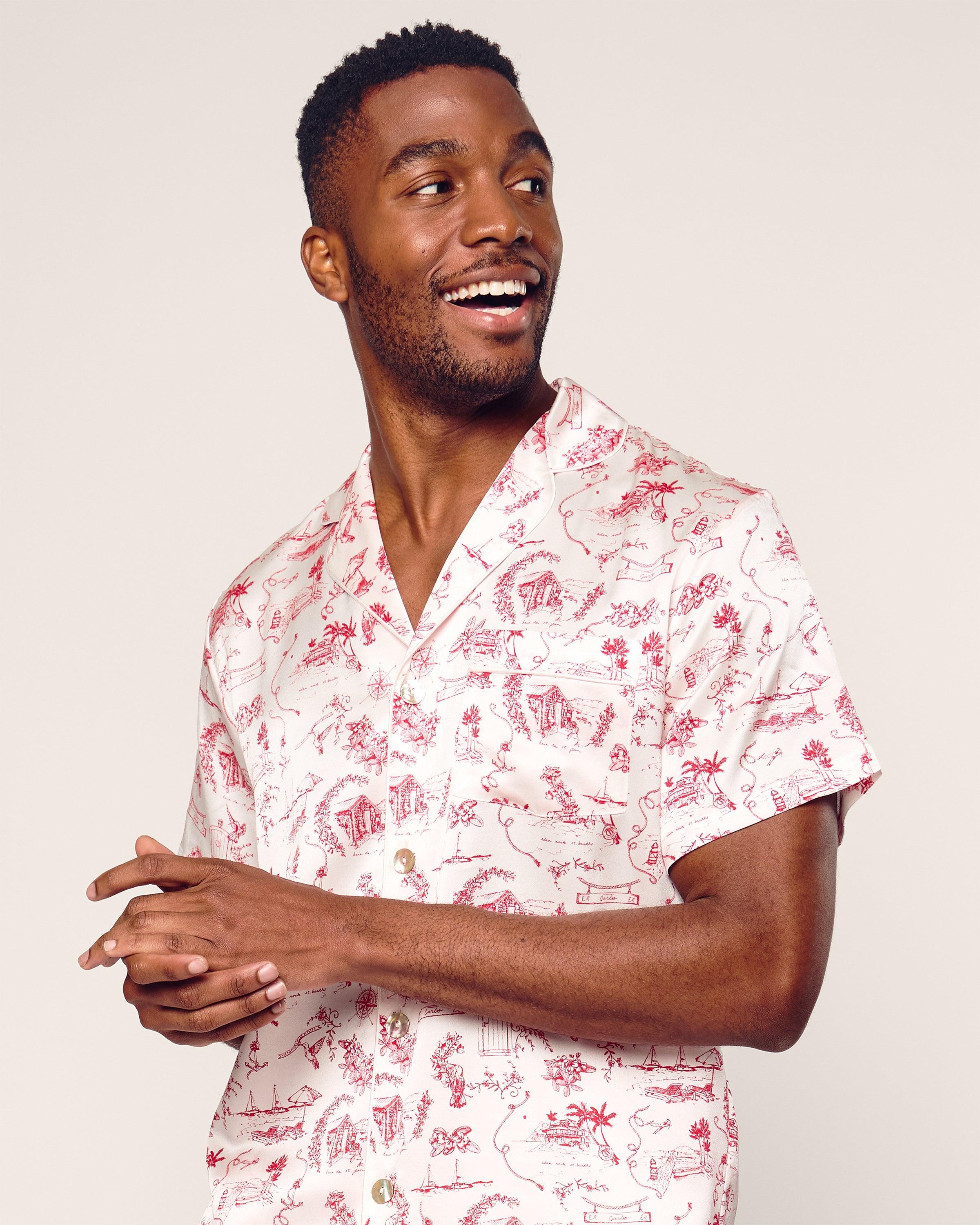 A person wearing a Petite Plume x Eden Rock Reverie Mens Silk Short Set, featuring a white and red pattern on a short-sleeve shirt, smiles and looks to the side, capturing the relaxed vibe of St Barths against a light gray background.