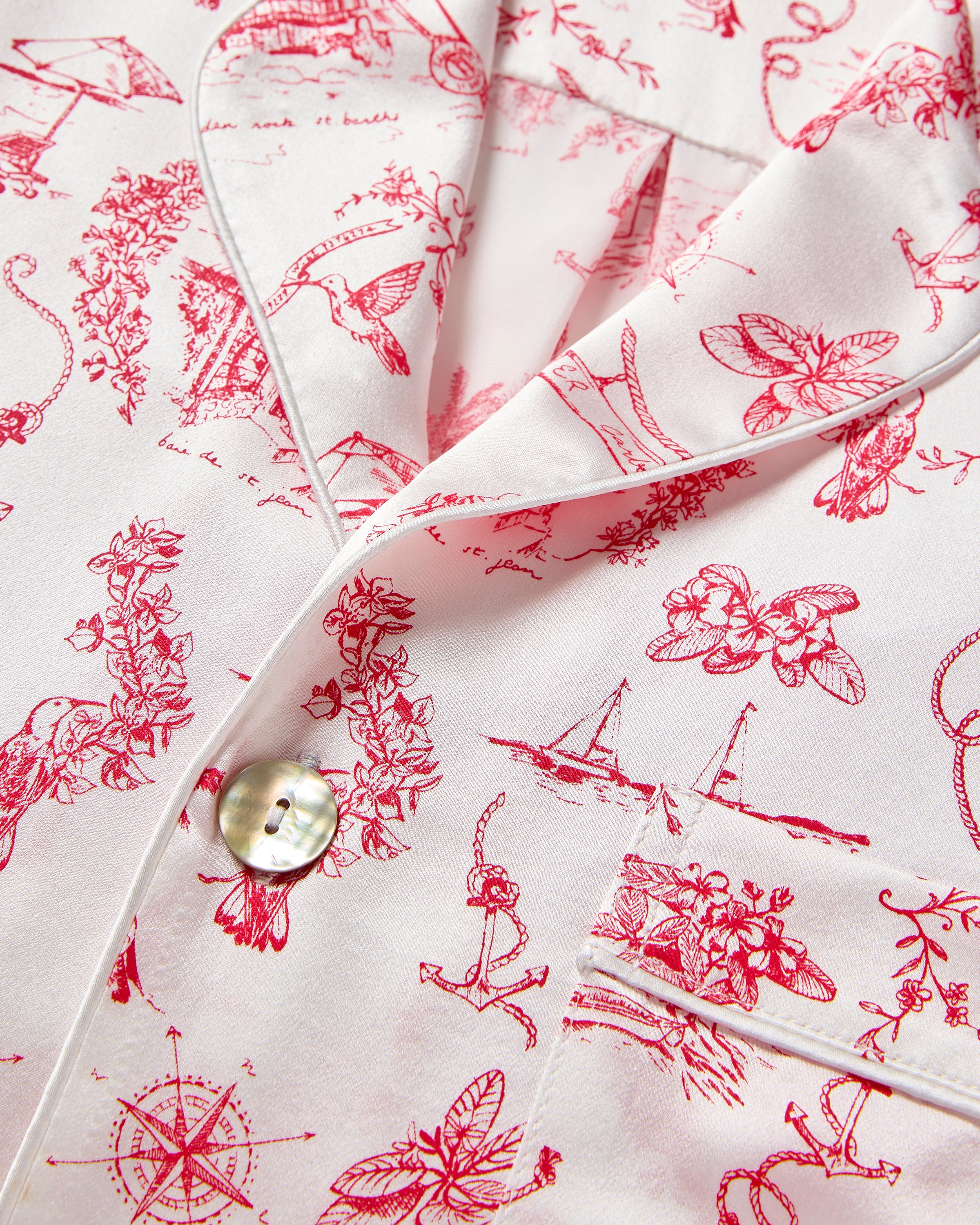 A close-up of the Petite Plume x Eden Rock Reverie Mens Silk Short Set shows mulberry silk fabric adorned with red nautical illustrations like anchors, sailboats, and compasses. A pearl-like button is visible above a pocket, part of this luxury sleepwear featuring piped edges.