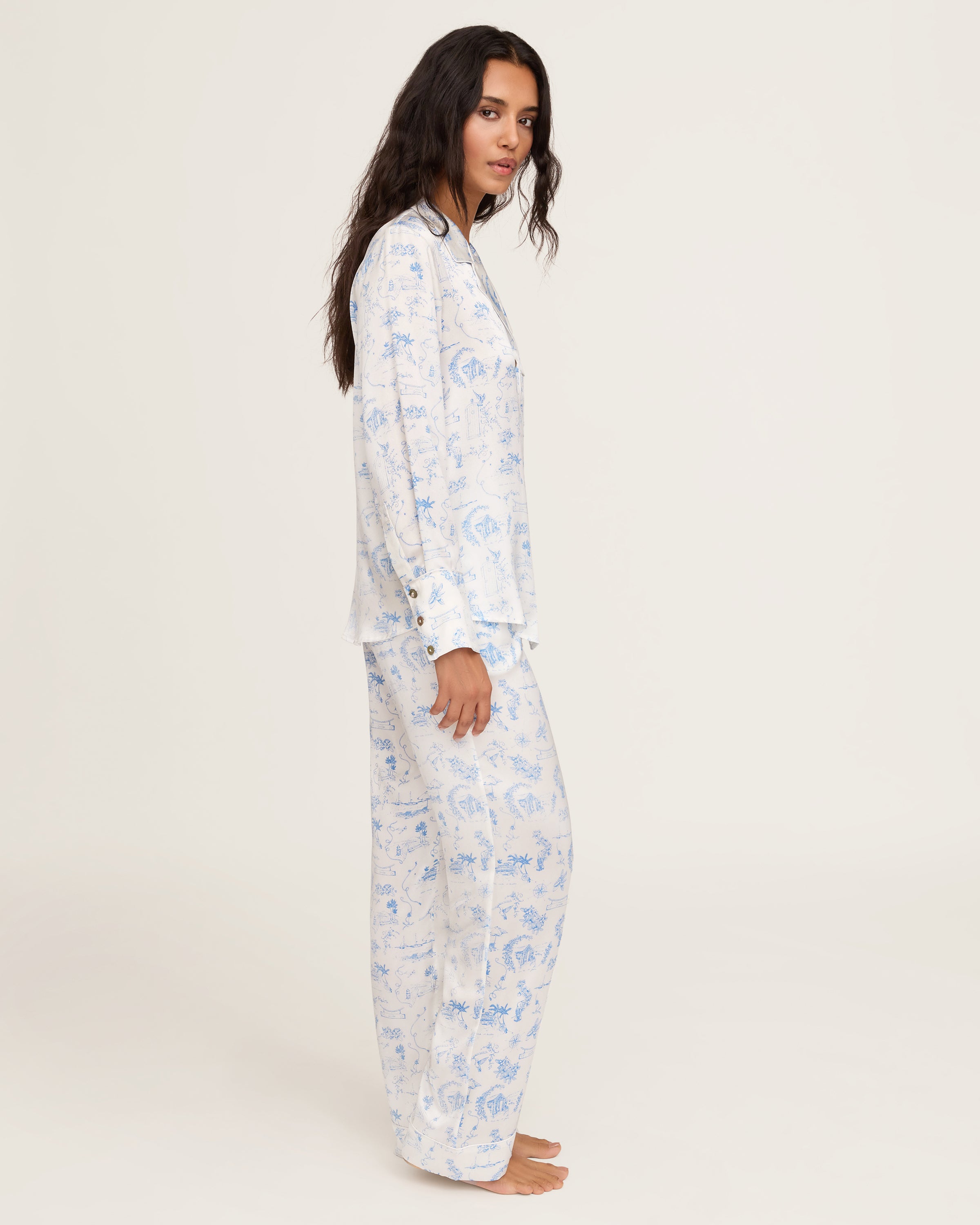 The Petite Plume x Eden Rock Blue Reverie Women's Silk Pajama Set features white pajamas with a blue floral and animal print, a long-sleeve collared button-up top, and matching pants with an elastic waistband.