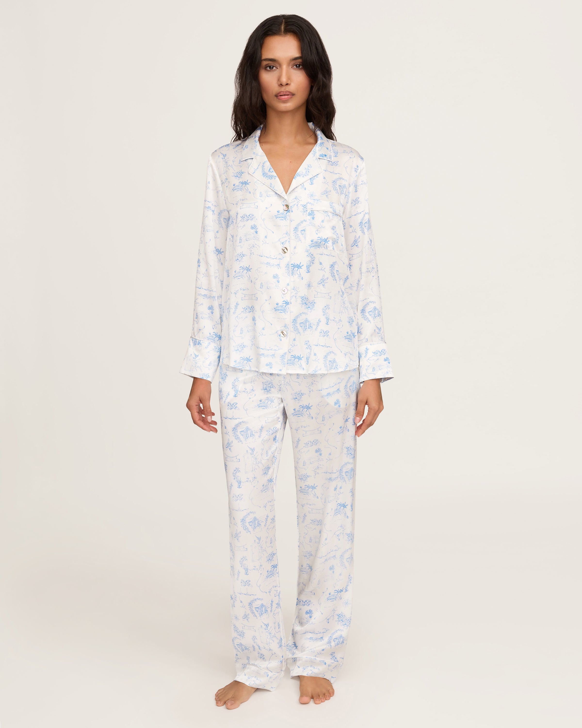 The Petite Plume x Eden Rock Blue Reverie Women's Silk Pajama Set features white pajamas with a blue floral and animal print, a long-sleeve collared button-up top, and matching pants with an elastic waistband.