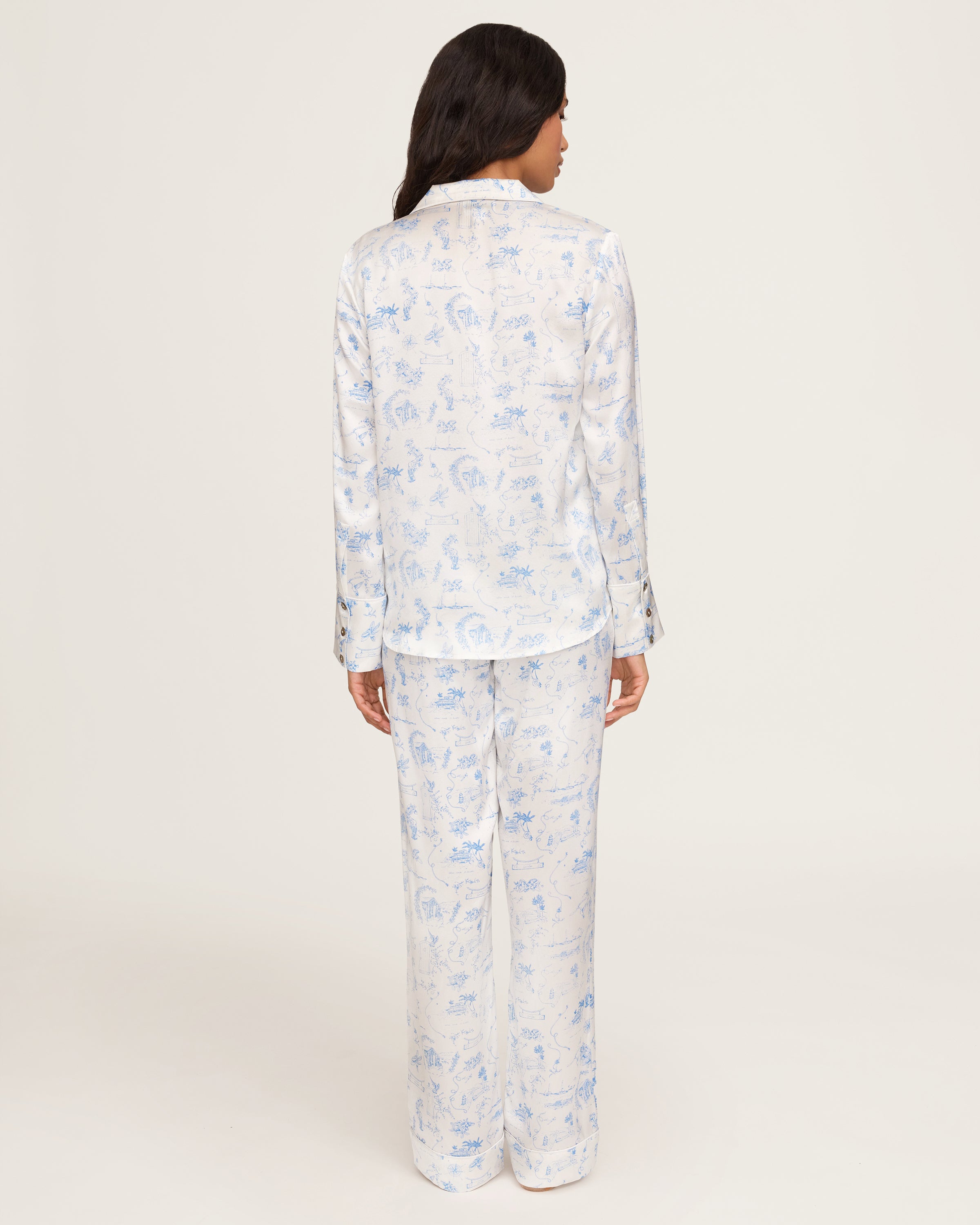 The Petite Plume x Eden Rock Blue Reverie Women's Silk Pajama Set features white pajamas with a blue floral and animal print, a long-sleeve collared button-up top, and matching pants with an elastic waistband.