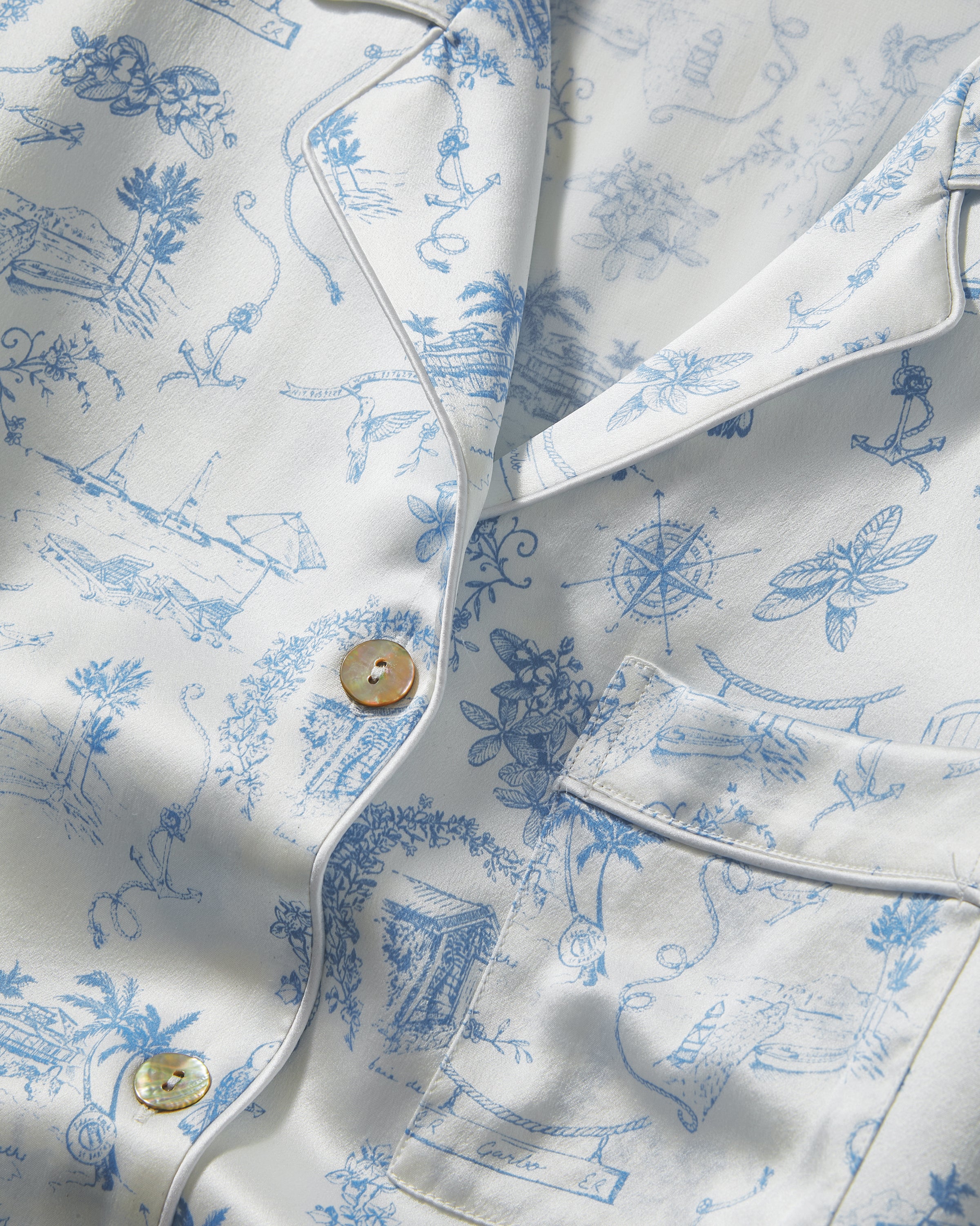 The Petite Plume x Eden Rock Blue Reverie Women's Silk Pajama Set features white pajamas with a blue floral and animal print, a long-sleeve collared button-up top, and matching pants with an elastic waistband.