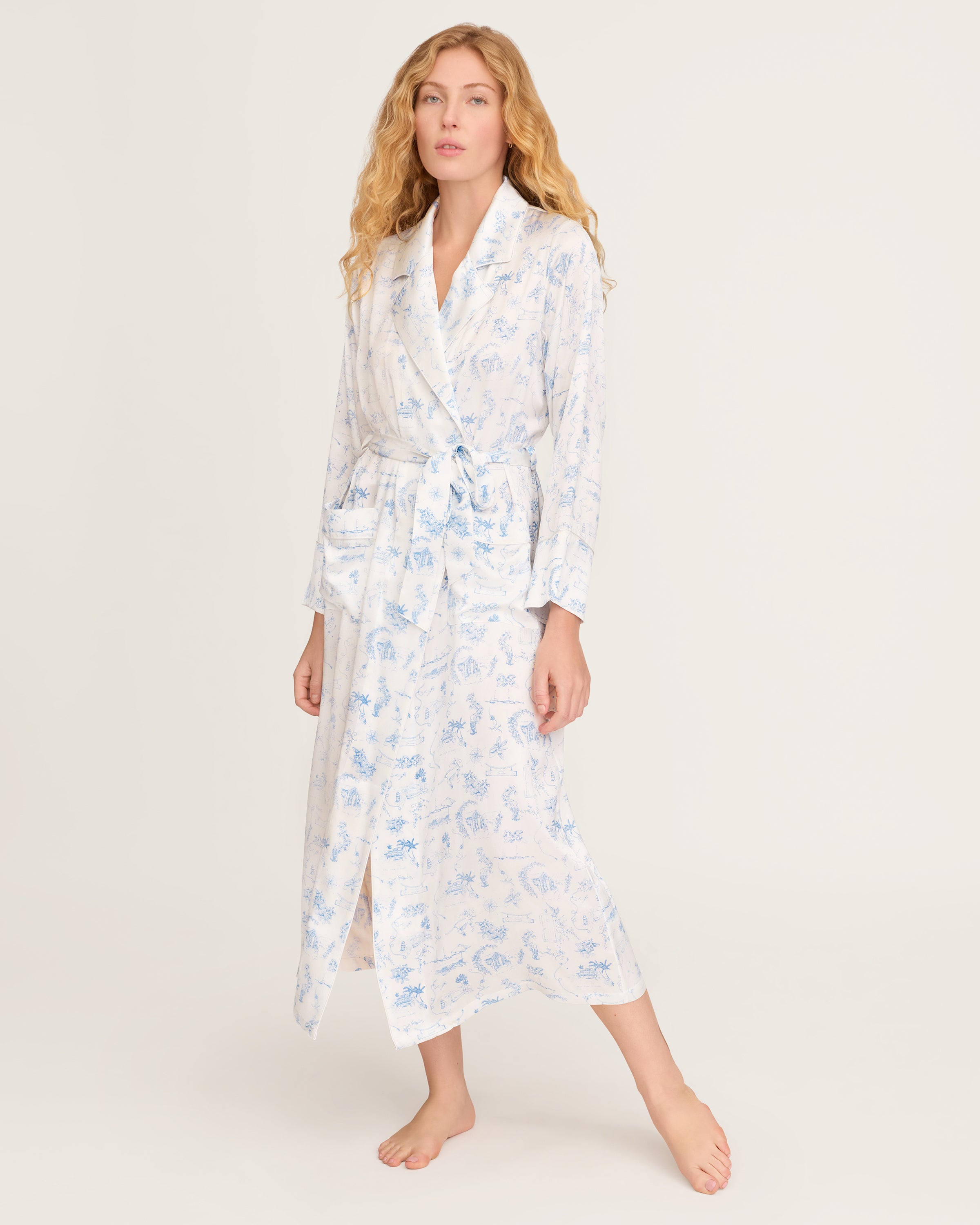 The Petite Plume x Eden Rock Blue Reverie Women's Silk Robe is a long, white robe with light blue toile print, notched collar, long sleeves, tie belt at the waist, and front patch pockets.