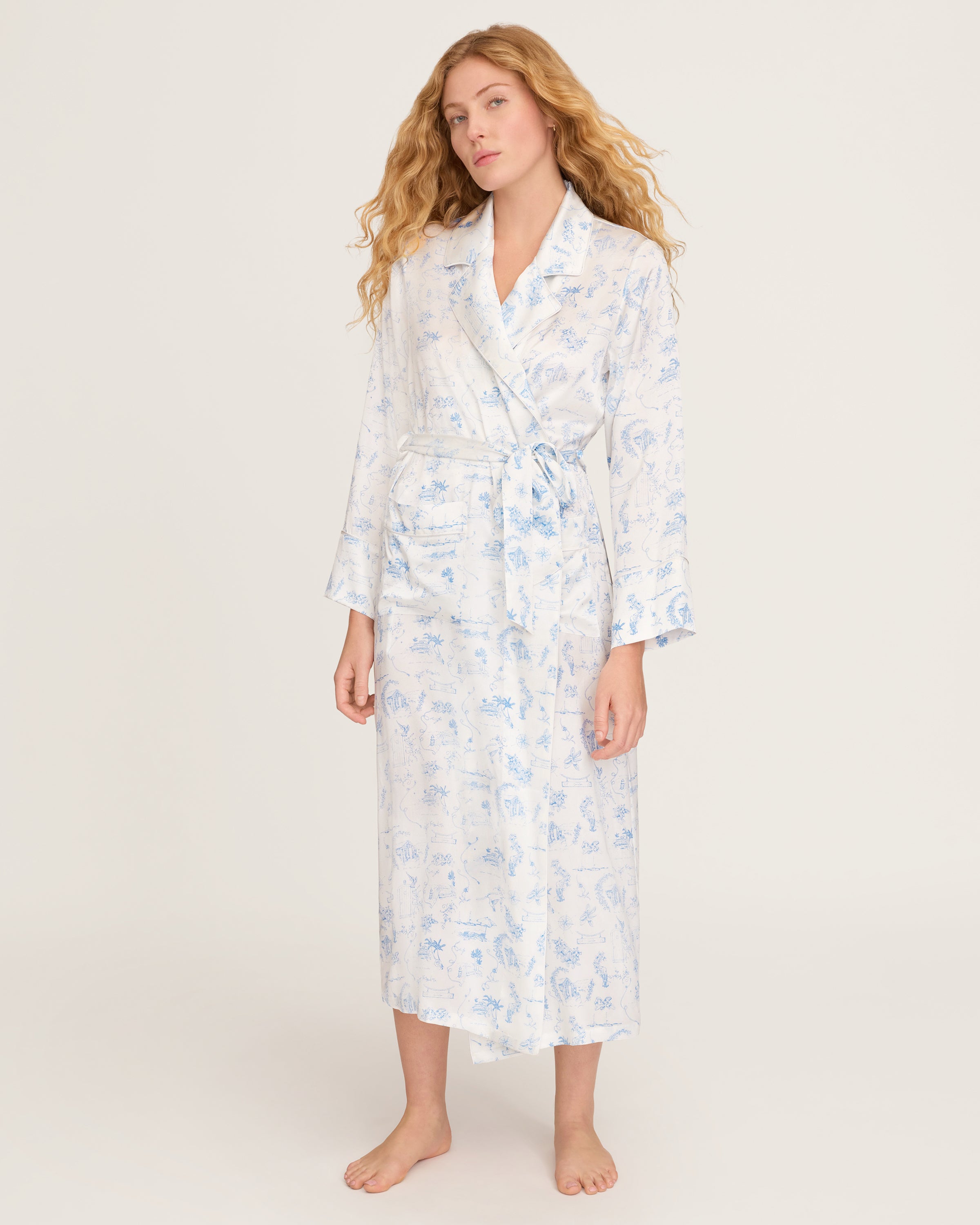 The Petite Plume x Eden Rock Blue Reverie Women's Silk Robe is a long, white robe with light blue toile print, notched collar, long sleeves, tie belt at the waist, and front patch pockets.