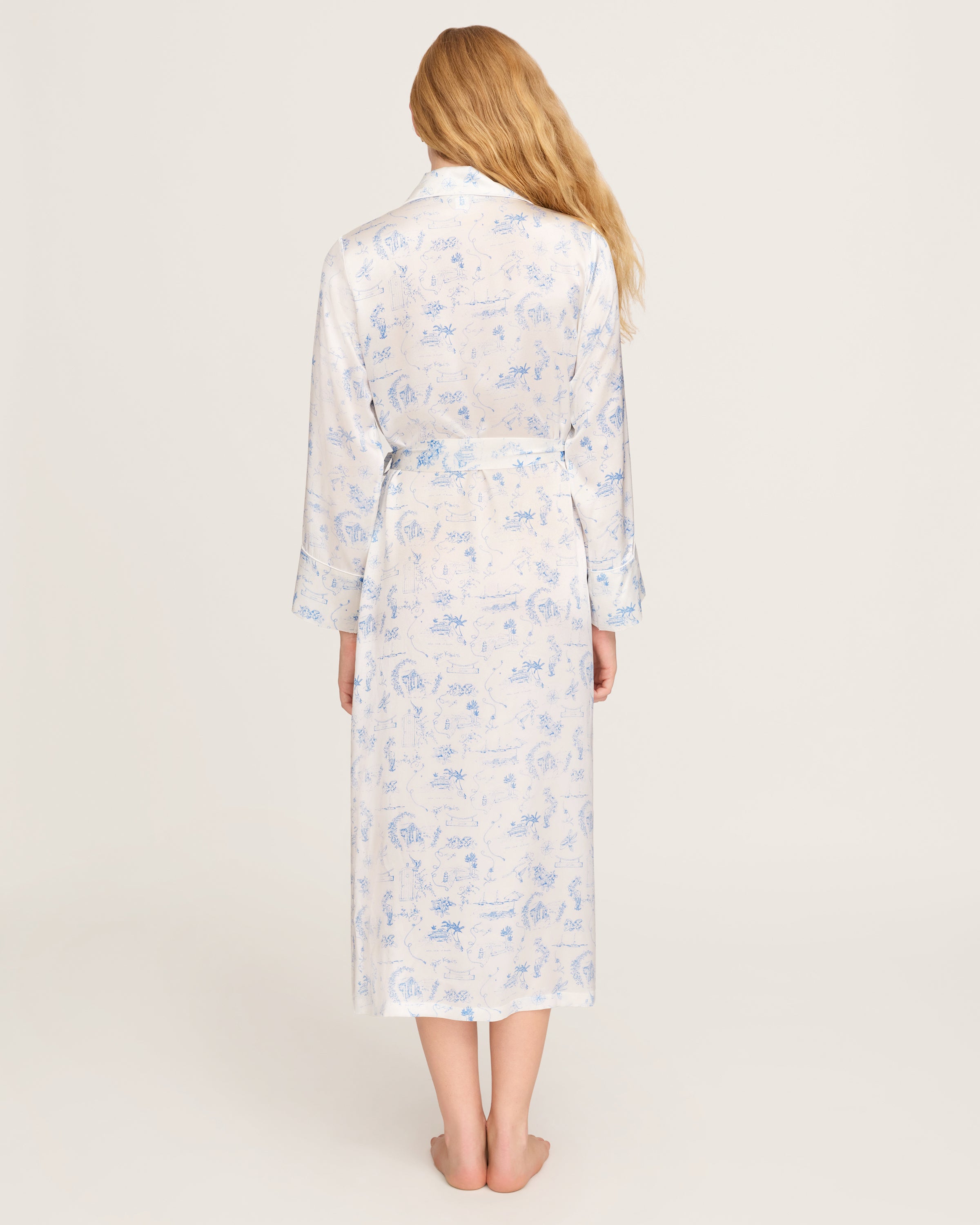 The Petite Plume x Eden Rock Blue Reverie Women's Silk Robe is a long, white robe with light blue toile print, notched collar, long sleeves, tie belt at the waist, and front patch pockets.