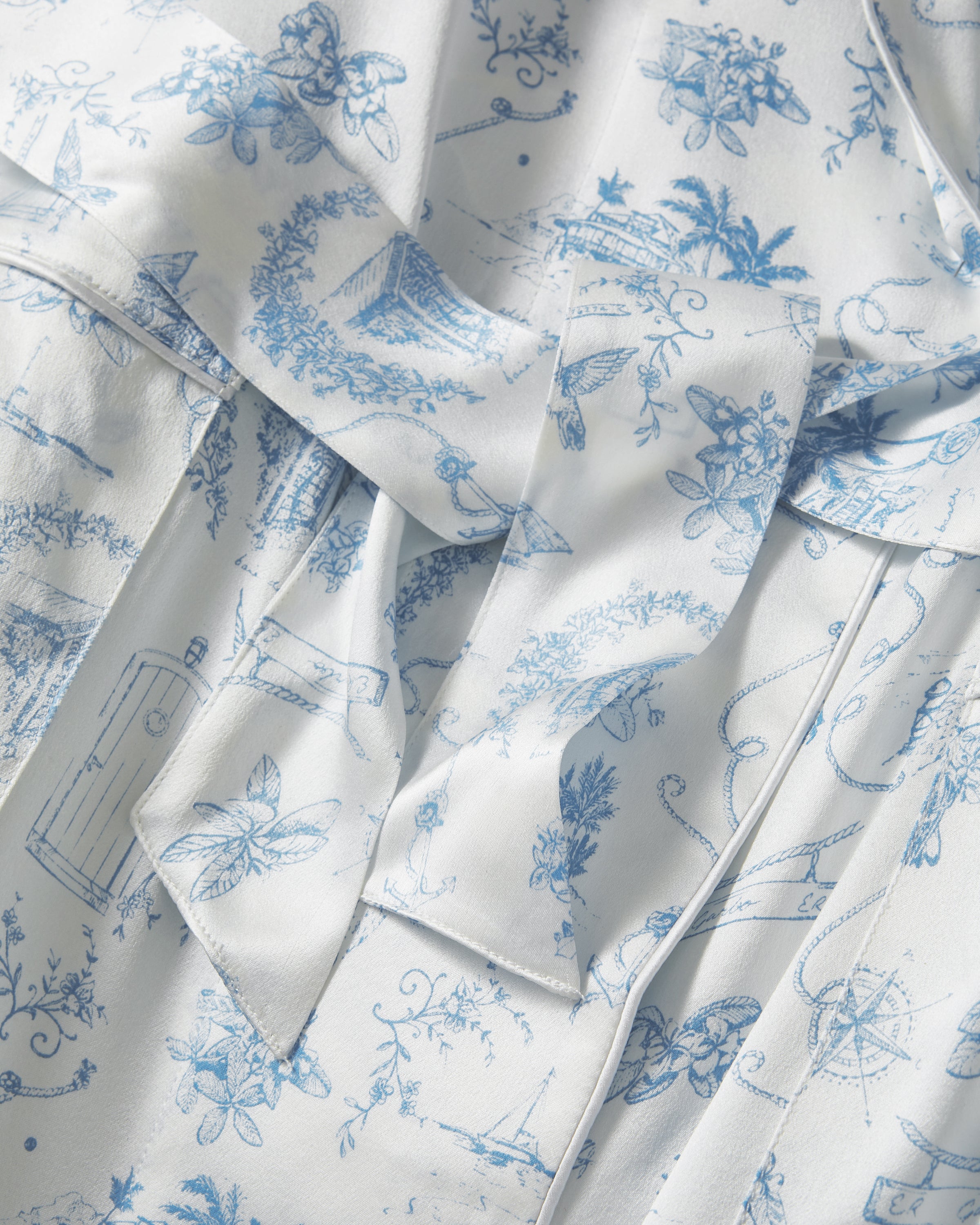 The Petite Plume x Eden Rock Blue Reverie Women's Silk Robe is a long, white robe with light blue toile print, notched collar, long sleeves, tie belt at the waist, and front patch pockets.
