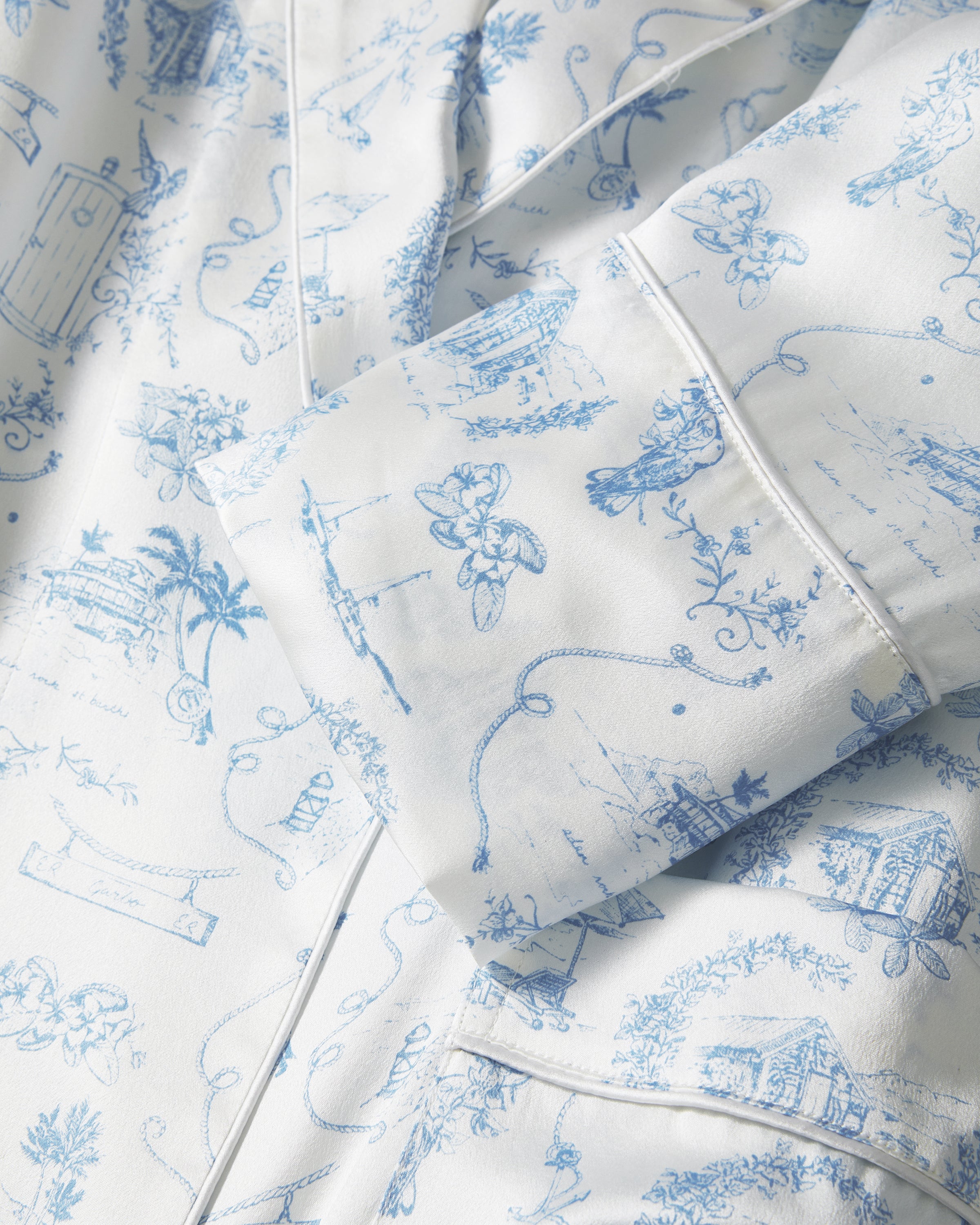 The Petite Plume x Eden Rock Blue Reverie Women's Silk Robe is a long, white robe with light blue toile print, notched collar, long sleeves, tie belt at the waist, and front patch pockets.