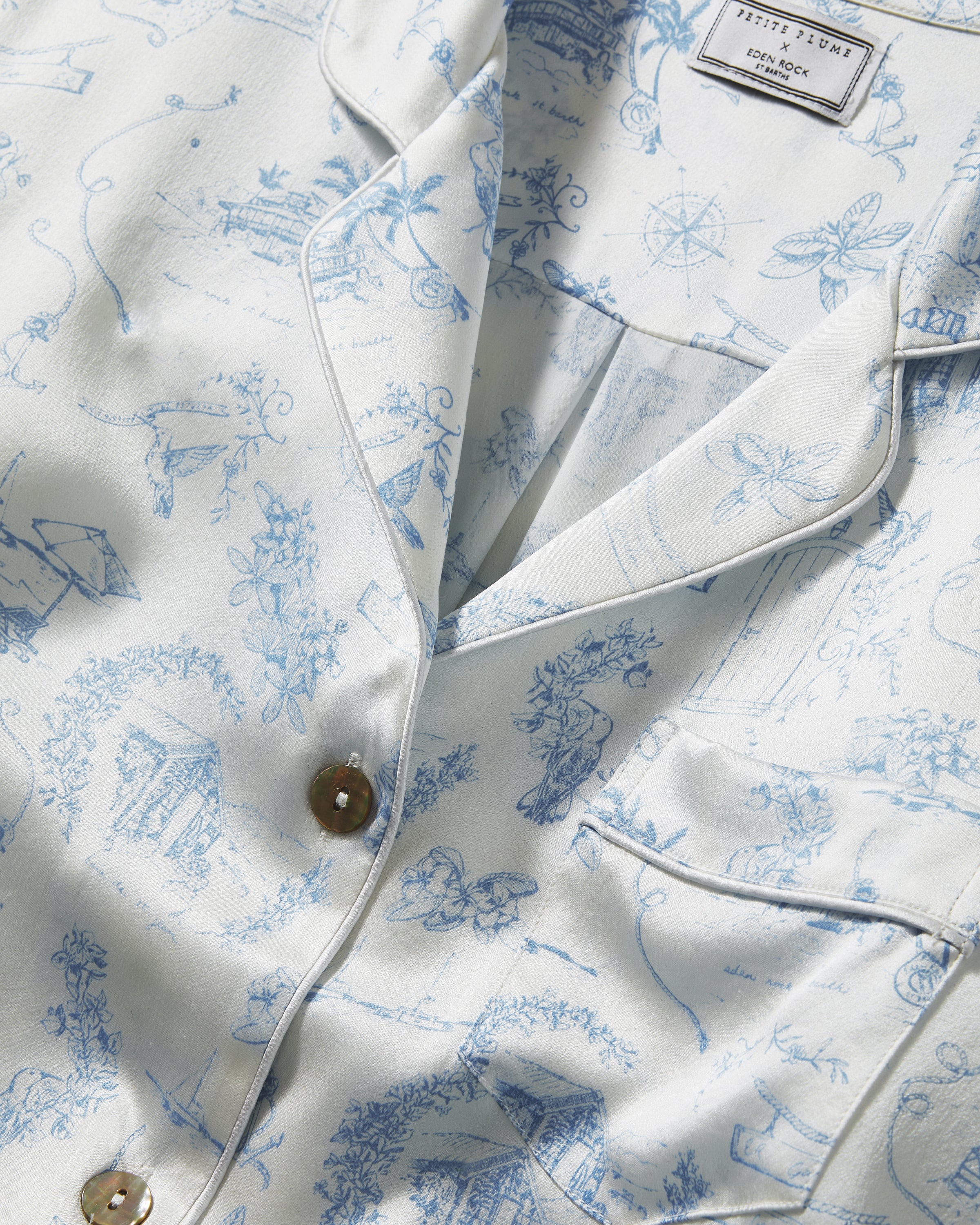 The Petite Plume x Eden Rock Blue Reverie Women's Silk Short Set features a white base with light blue nature-inspired prints, a short-sleeve button-up shirt with chest pocket, and drawstring waist shorts.