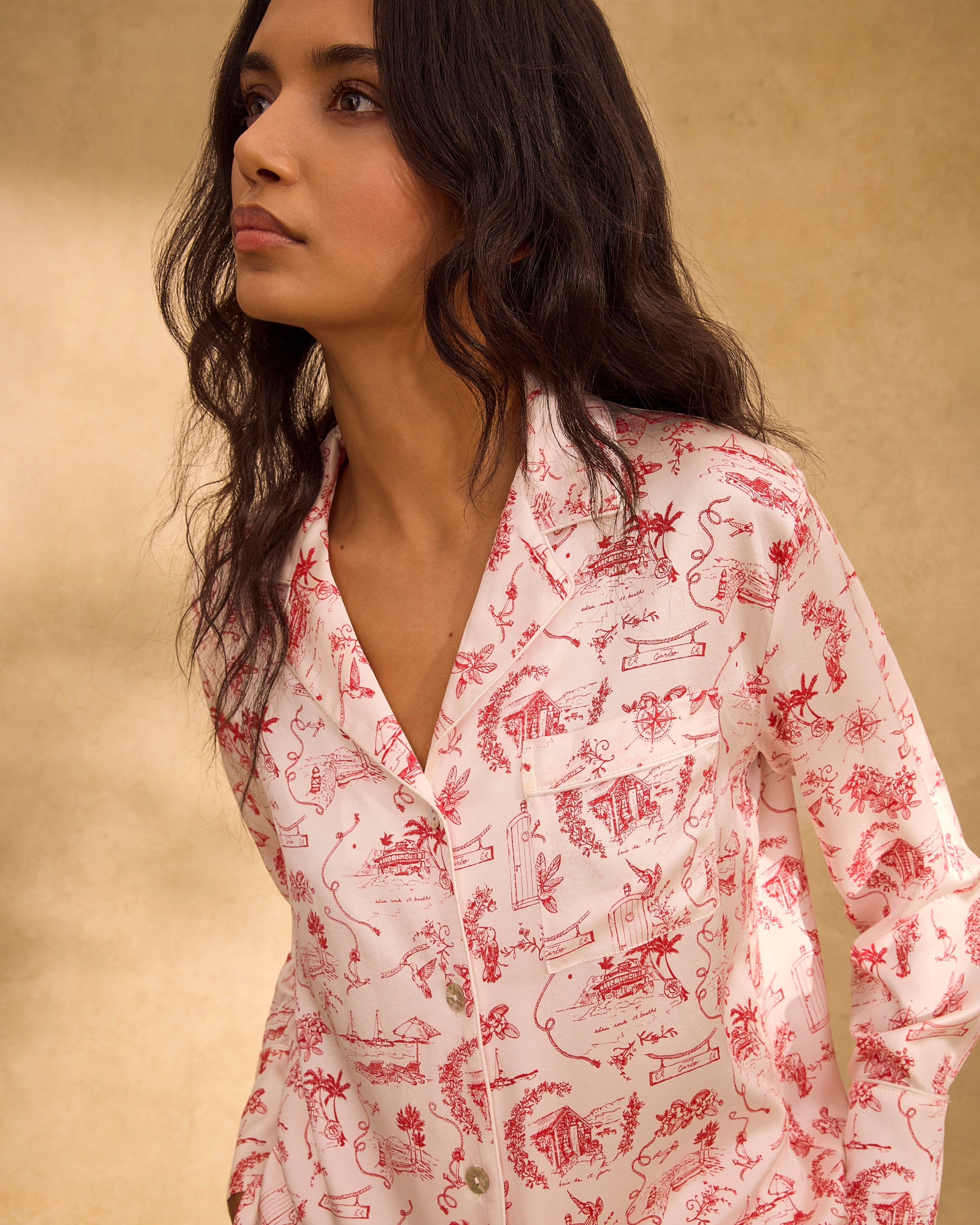 The Petite Plume x Eden Rock Reverie Women's Silk Pajama Set features a long-sleeve button-up top and pants with a white background and red toile print of scenic and animal motifs.