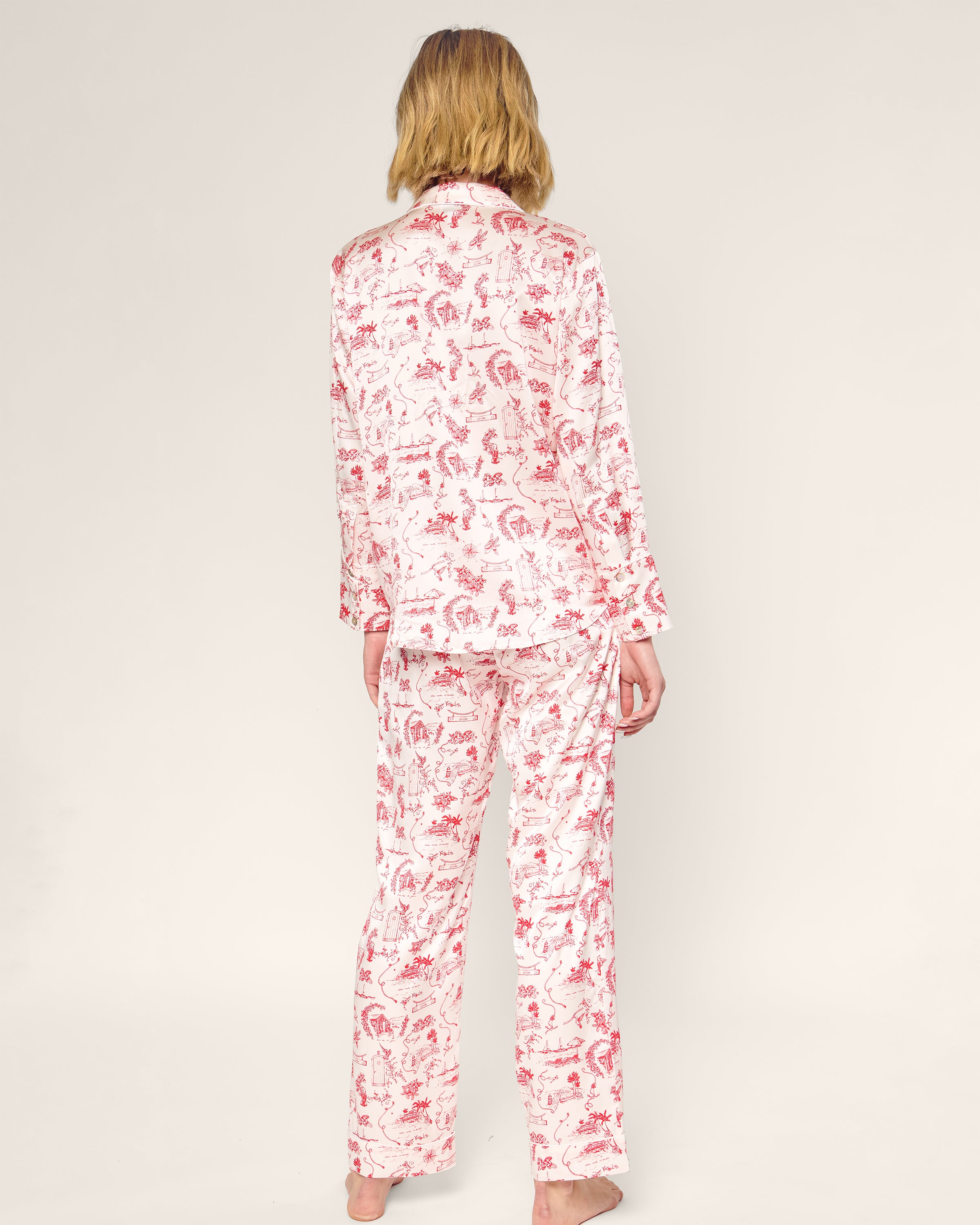 The Petite Plume x Eden Rock Reverie Women's Silk Pajama Set features a long-sleeve button-up top and pants with a white background and red toile print of scenic and animal motifs.