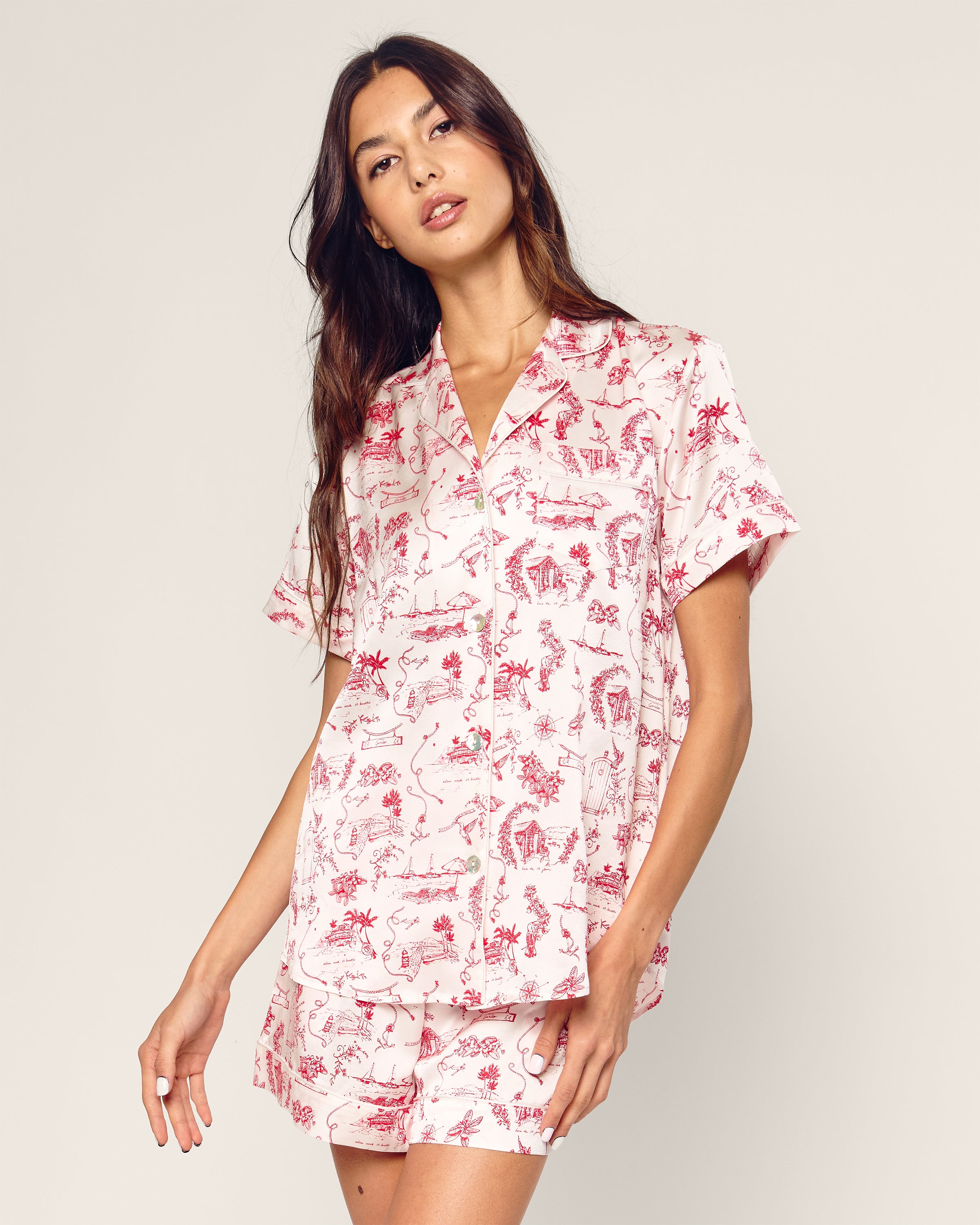 Wearing the Petite Plume x Eden Rock Reverie Womens Silk Short Set, featuring a white background with red toile print and a short-sleeved button-up shirt with shorts, the individual has long dark hair and stands against an off-white backdrop, exuding effortless elegance.