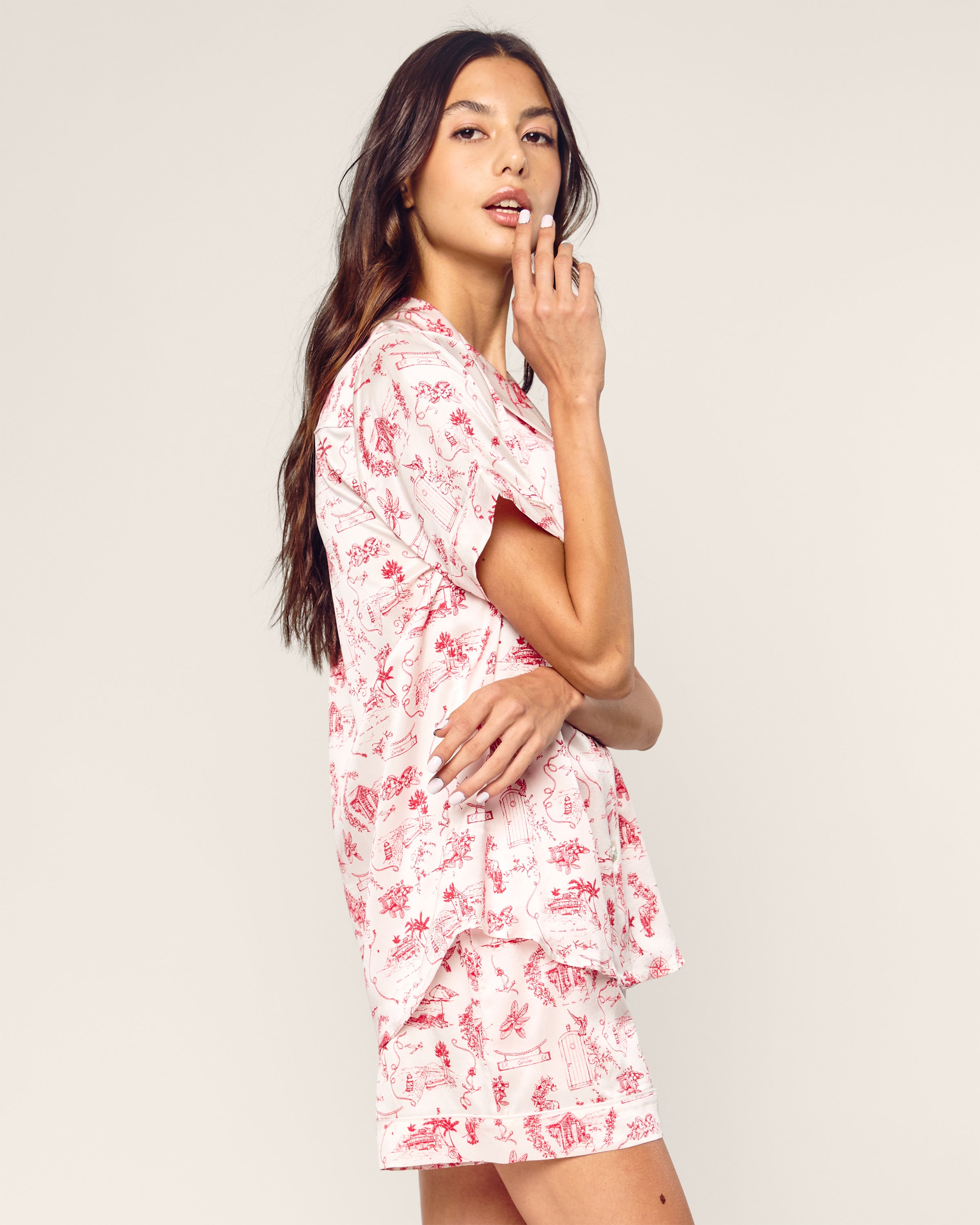 A person with long hair is wearing the luxury Petite Plume x Eden Rock Reverie Womens Silk Short Set in pink and white. Made from exquisite mulberry silk, they stand sideways, gently touching their lips against a light-colored background.