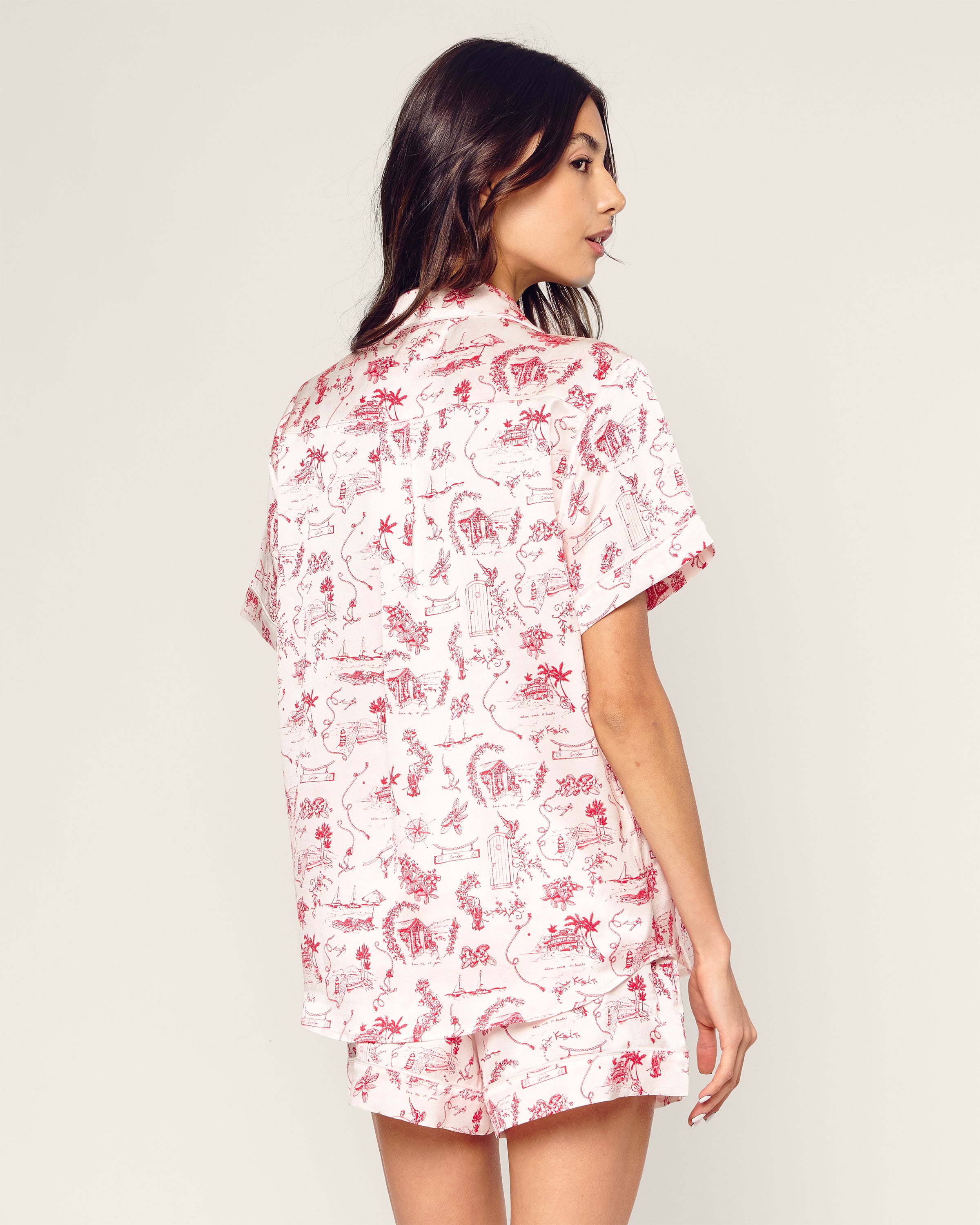 A woman with long dark hair stands turned slightly away, wearing the Petite Plume x Eden Rock Reverie Womens Silk Short Set in mulberry silk with a red and white toile design. The backdrop is a plain, light hue, capturing the serene elegance of St Barths.