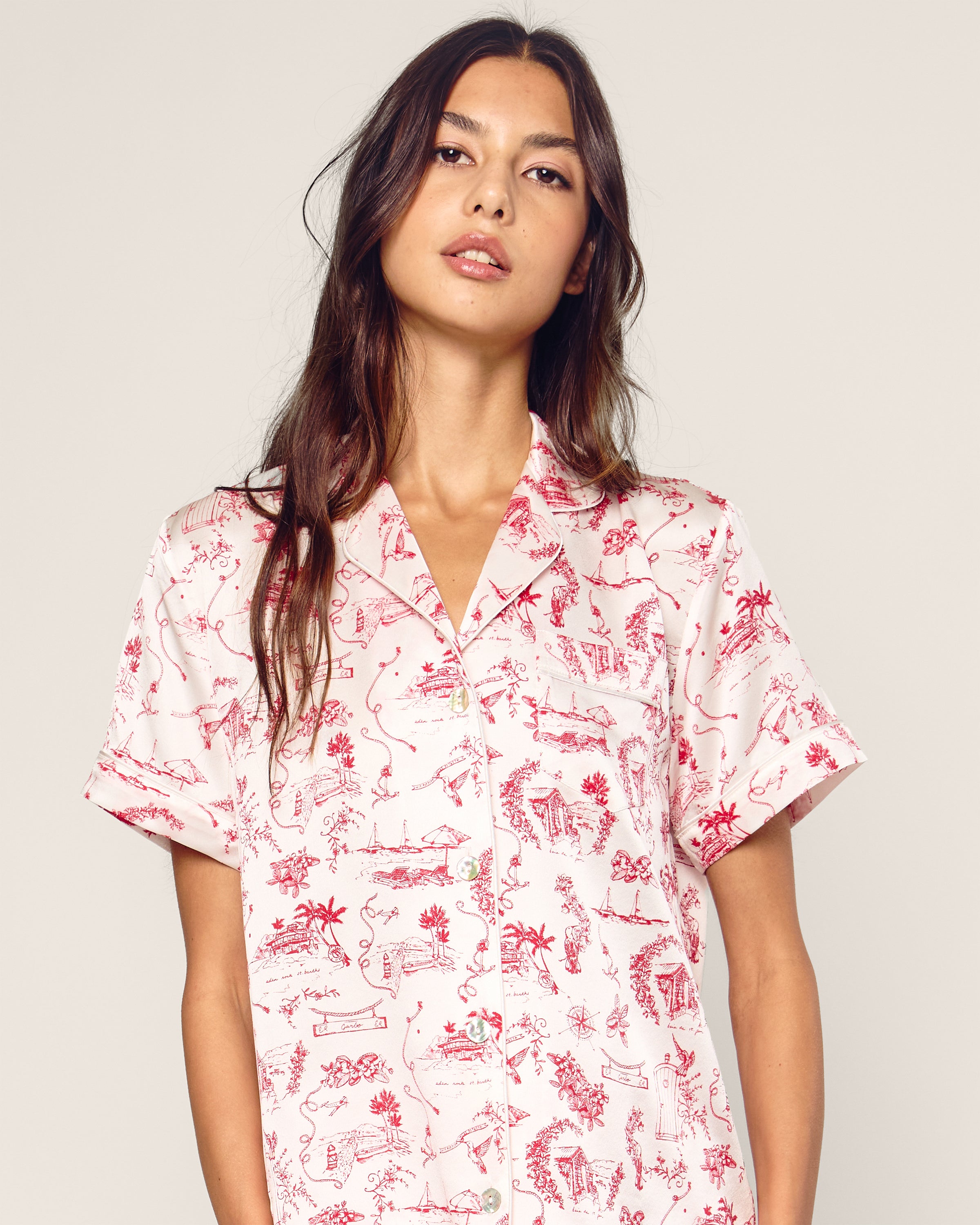 A person with long, dark hair wears the Petite Plume x Eden Rock Reverie Womens Silk Short Set, featuring a light-colored short-sleeve shirt with red patterns. They stand against a neutral background, looking slightly to the side and evoking an air of relaxed elegance reminiscent of St Barths.