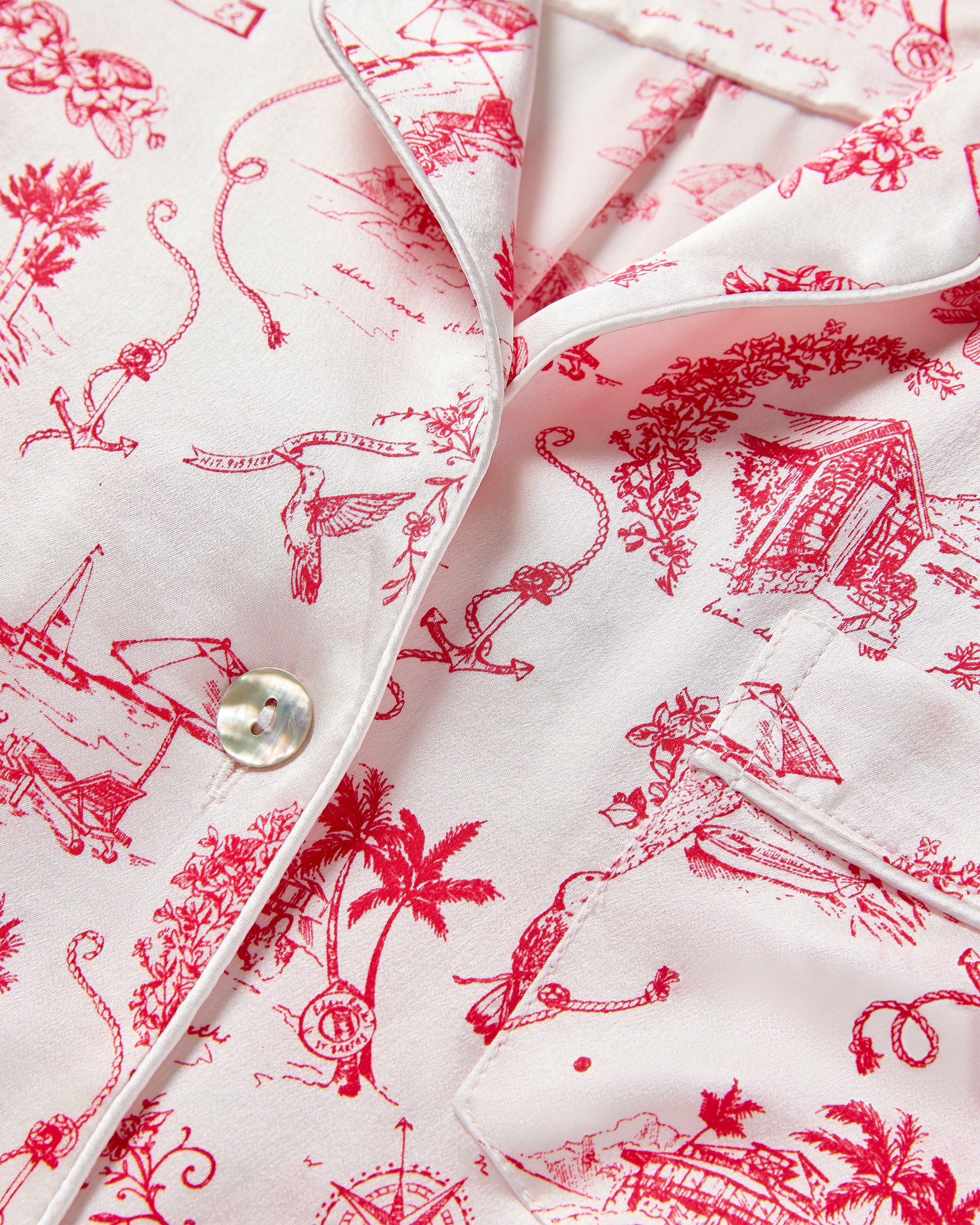 A close-up of the Petite Plume x Eden Rock Reverie Womens Silk Short Set shows luxurious white mulberry silk fabric with red tropical illustrations of palm trees, monkeys, birds, and huts. This vintage-style piece includes a button and a piped pocket edge, reminiscent of St Barths.