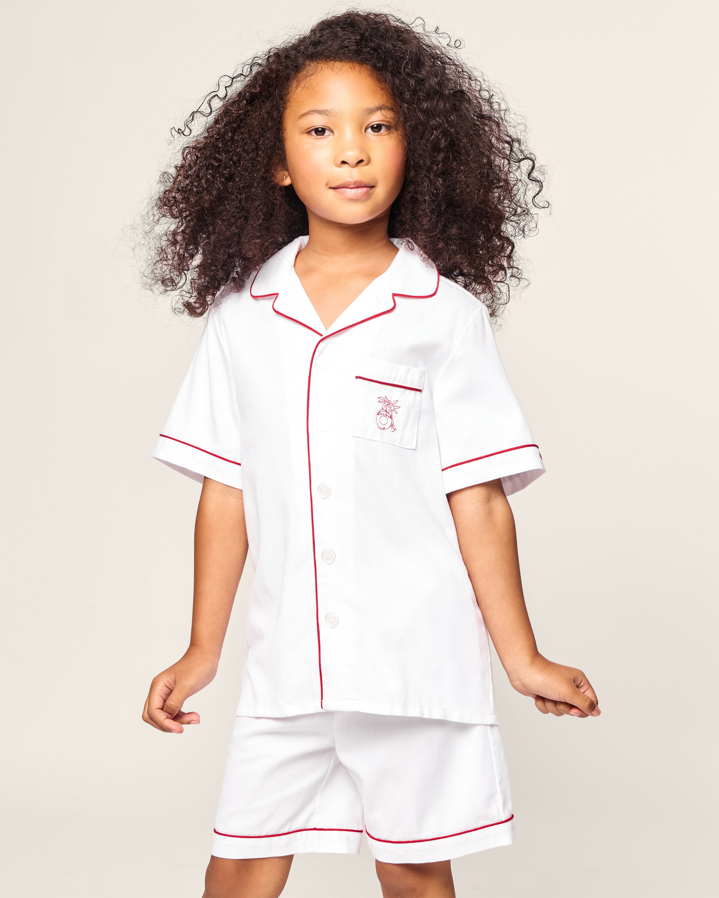 A young child with curly hair confidently wears the Petite Plume x Eden Rock St Barth Kids White Twill Short Set, luxurious sleepwear featuring white pajamas with red piping and a pocket with an embroidered design from the limited-edition collection, set against a plain background.