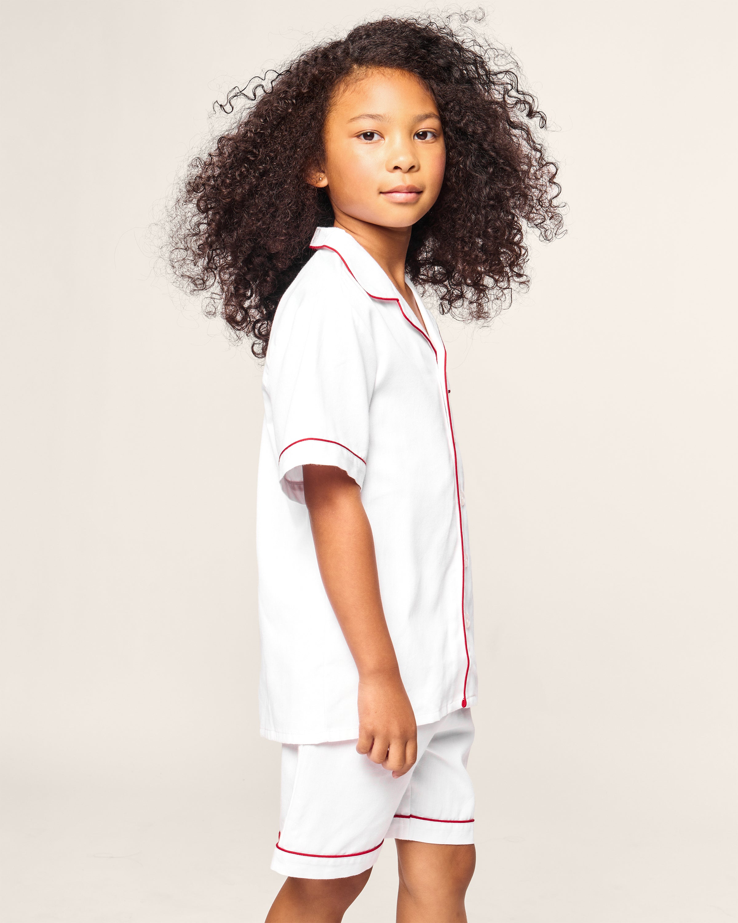A child with long curly hair stands sideways, wearing the limited-edition Petite Plume x Eden Rock St Barth Kids White Twill Short Set, evoking luxurious sleepwear. The background is plain light gray.
