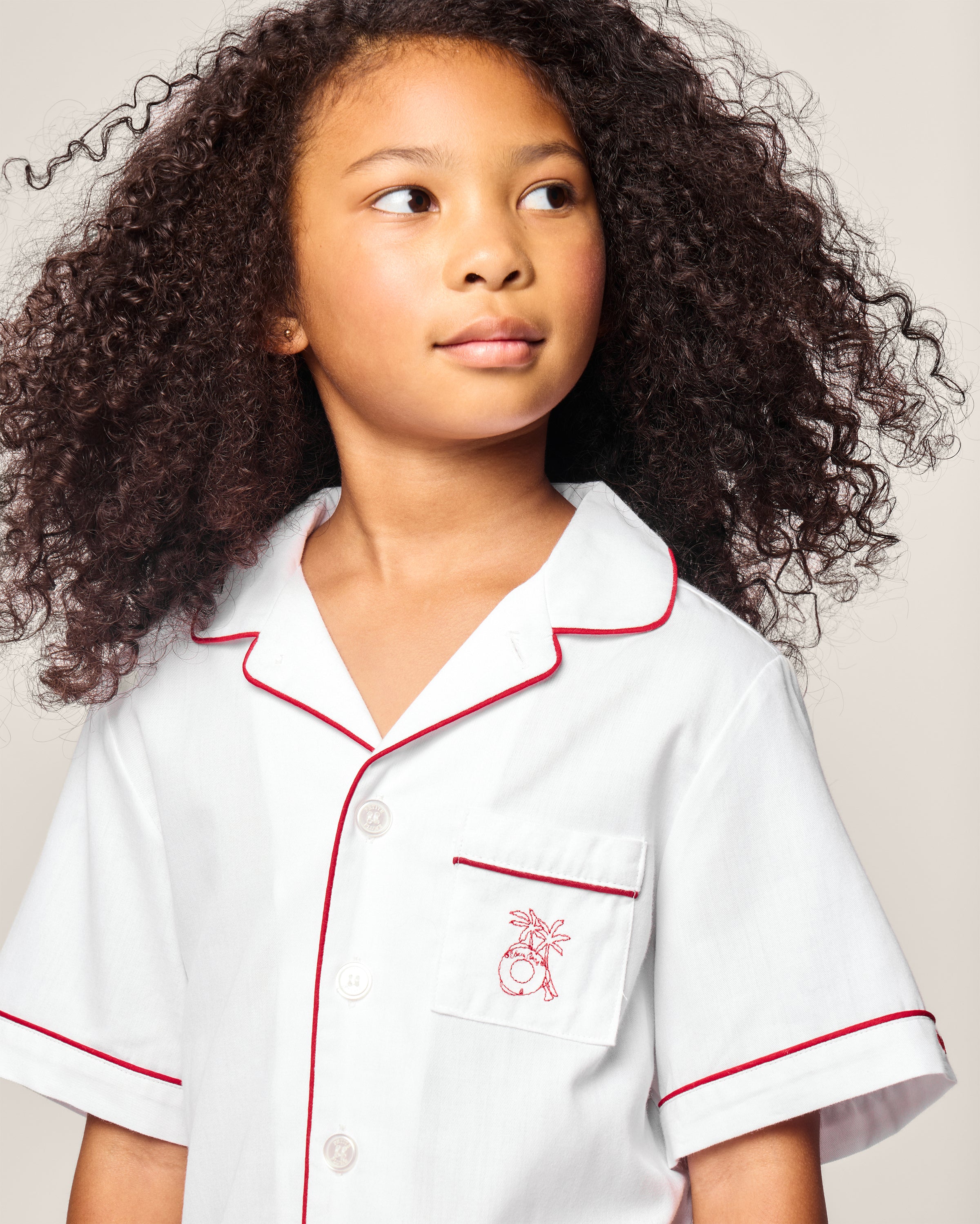 A young girl with curly hair models the Petite Plume x Eden Rock St Barth Kids White Twill Short Set, featuring red piping and a small red embroidered design on the pocket. She gazes to the side neutrally against a light background, embodying relaxed elegance.
