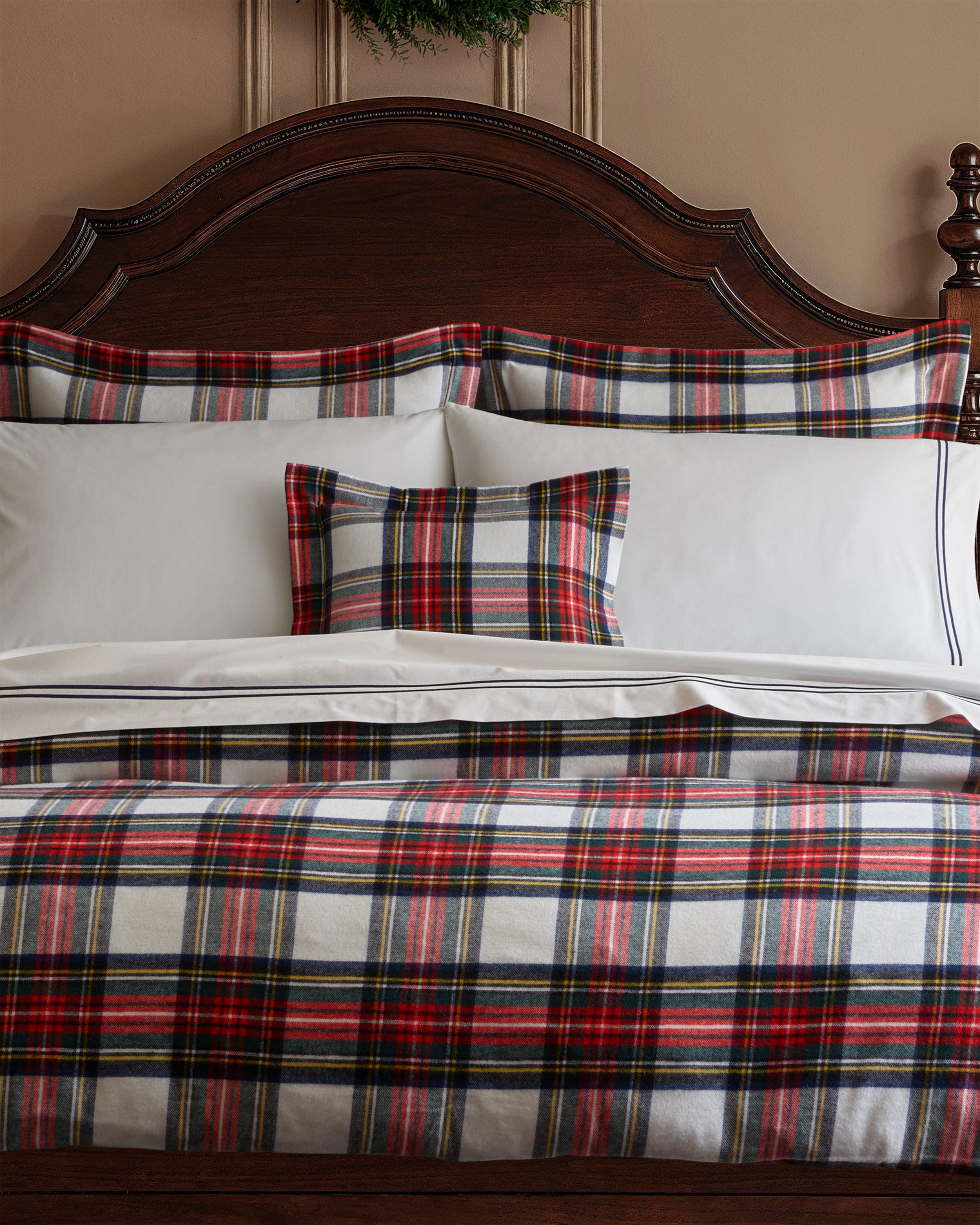 A bedroom with a dark wooden headboard features a bed adorned in Petite Plumes Flannel Euro Sham in Balmoral Tartan, highlighting red, white, blue, and green plaid on the comforter and matching pillow sham. The taupe walls enhance the decor, as a small green wreath hangs above the bed.