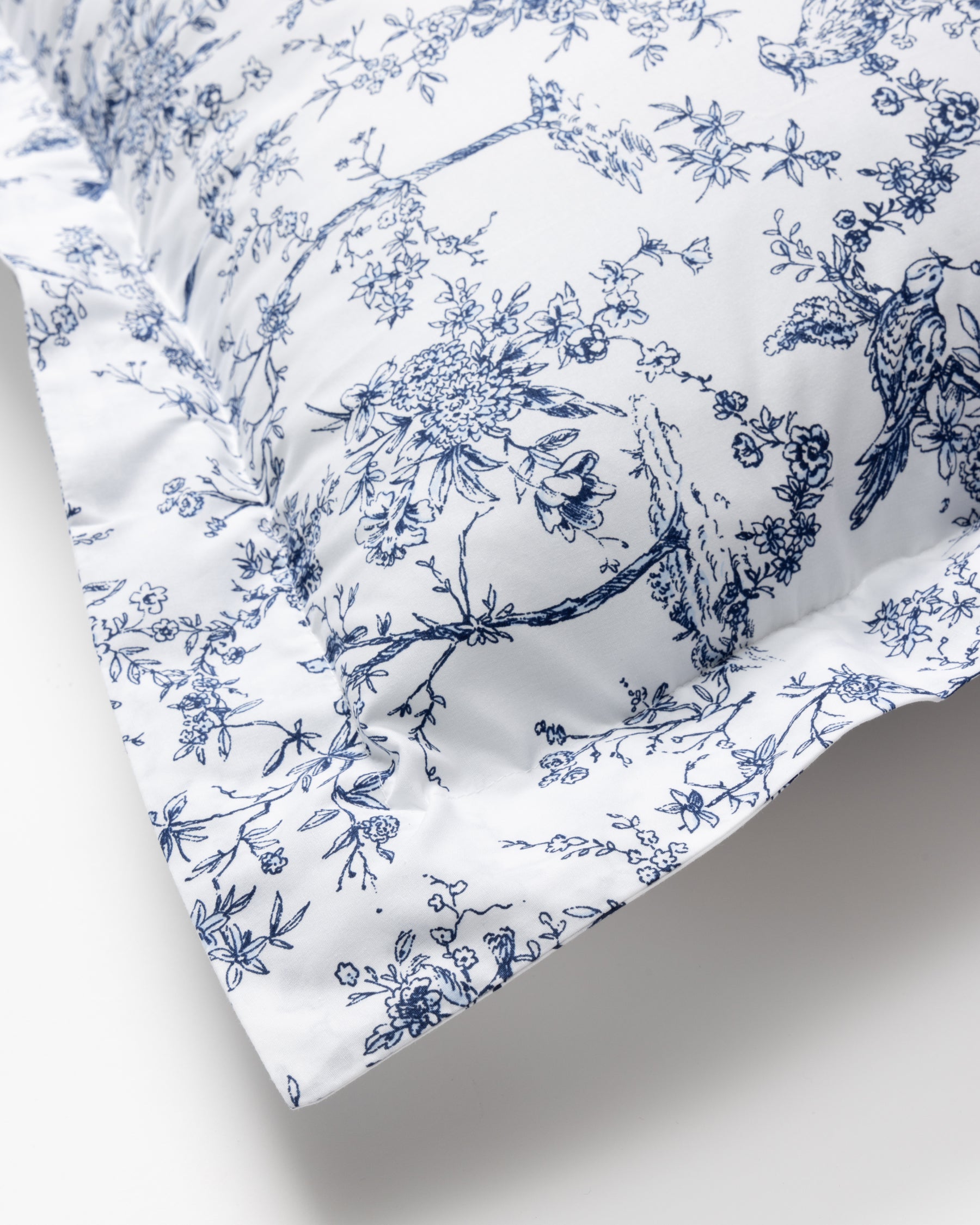 A close-up of the Luxe Premium Cotton Euro Sham in Timeless Toile by Petite Plume reveals its intricate blue floral and bird motifs on percale fabric, offering a delicate and elegant appearance with its prominently displayed corner.