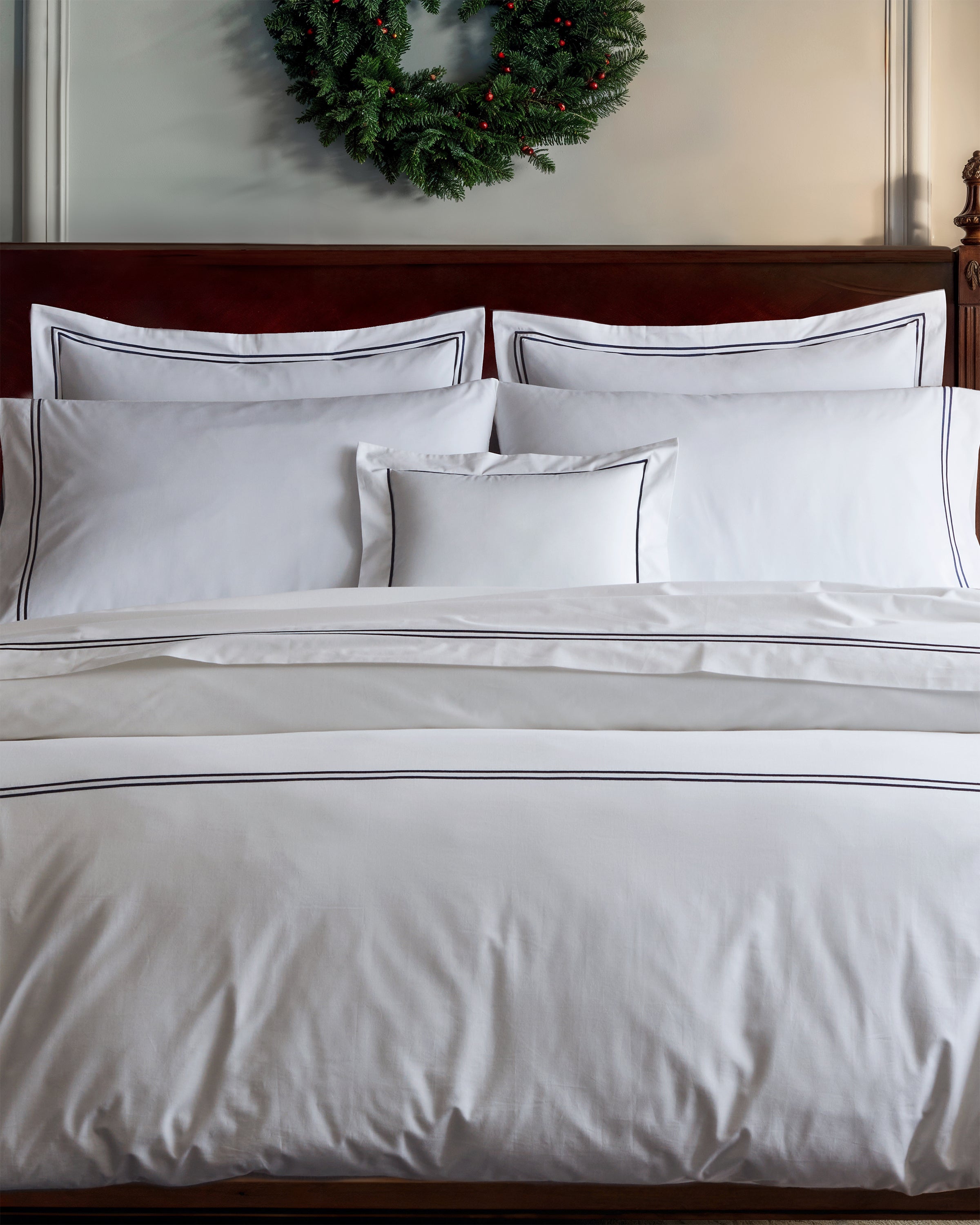 A neatly made bed features a Luxe Premium Percale Euro Sham in White with Navy by Petite Plume, centered as a small cushion. Above the wooden headboard, a festive green wreath adds coziness to the room.