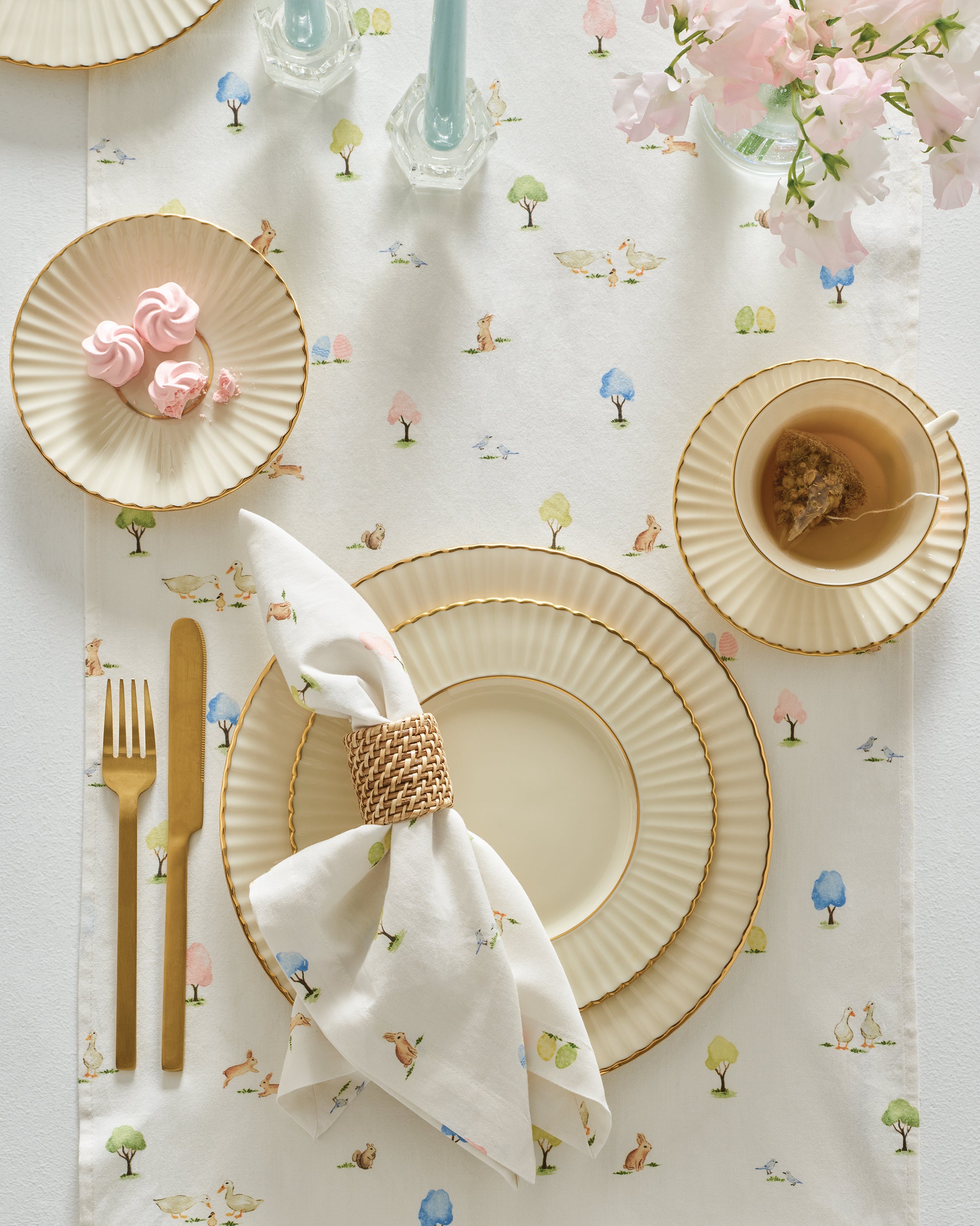A festive tablescape showcases Petite Plumes Easter Gardens Table Linen, featuring a whimsical tree and animal design. It is complemented by fluted-edge plates, a gold cutlery set, pink meringues on a small plate, a teacup, and a white napkin in a wicker ring. Pink flowers add to the charm.