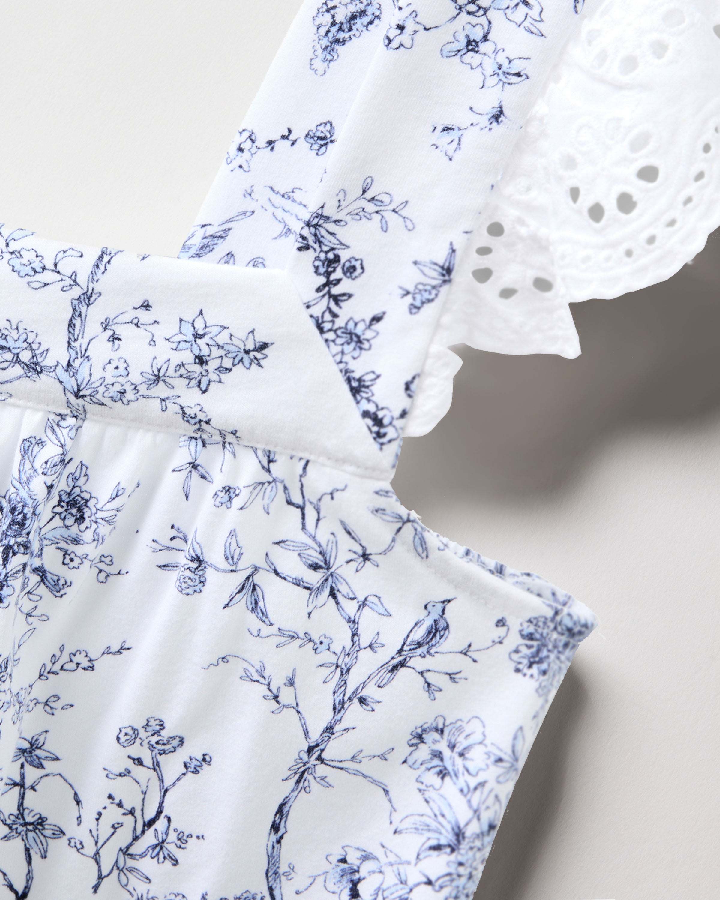 The Womens Pima Clara Short Set from Petite Plumes Timeless Toile collection features a close-up of white fabric adorned with delicate blue floral and bird patterns, complete with intricate eyelet lace edges for a vintage and elegant look.