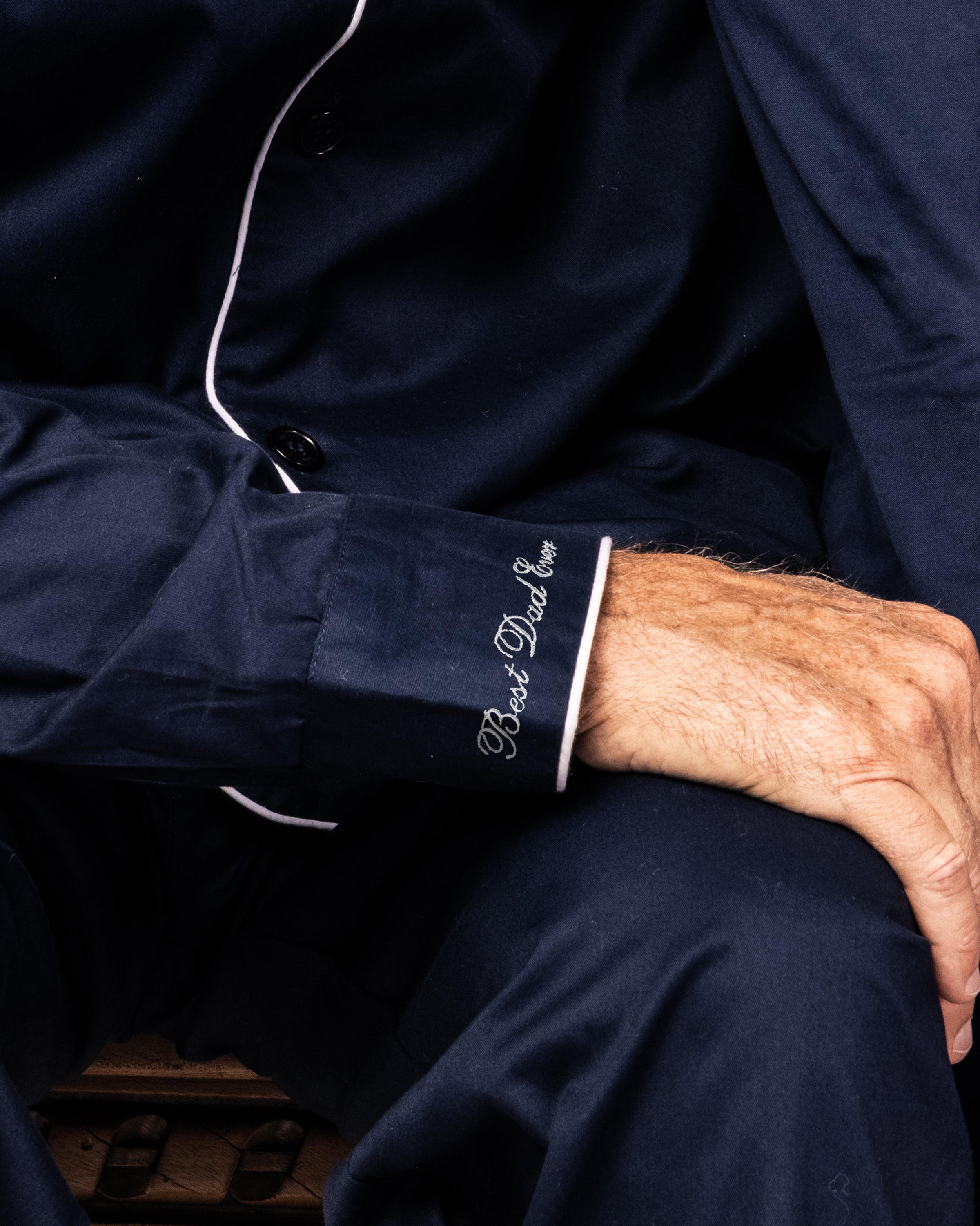 A person wears Petite Plumes Mens Twill Navy Pajama, featuring white piping and a Best Dad Ever embroidery on the cuff, with their hand resting on their knee.