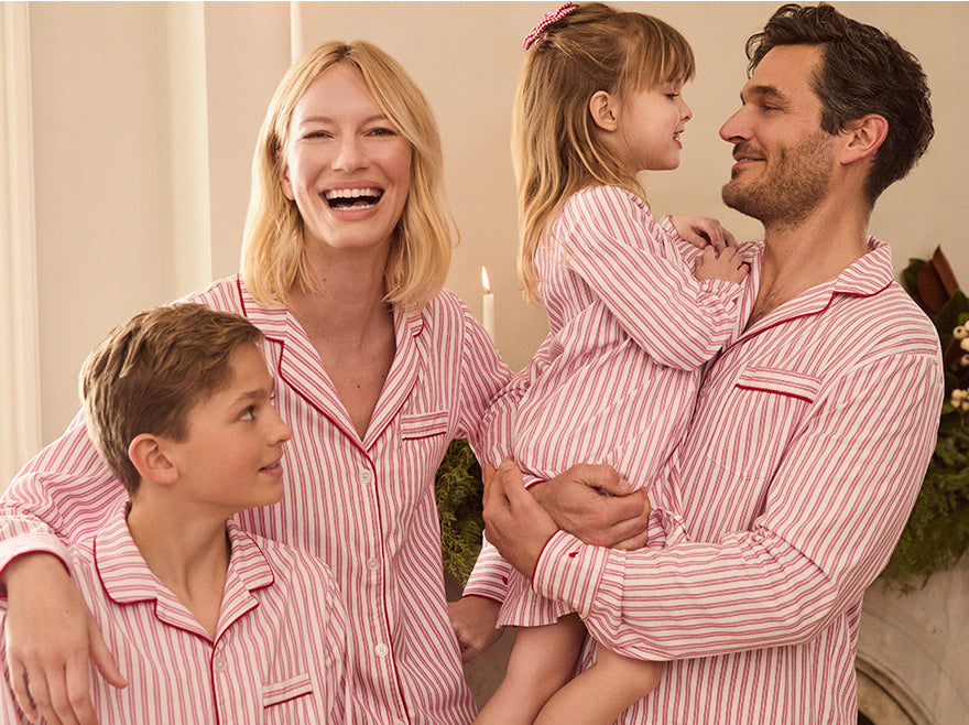 A smiling family of four, all wearing matching red-and-white striped pajamas, stands together indoors. The father holds a young girl, while the mother and a boy stand close by, all appearing happy and cheerful.