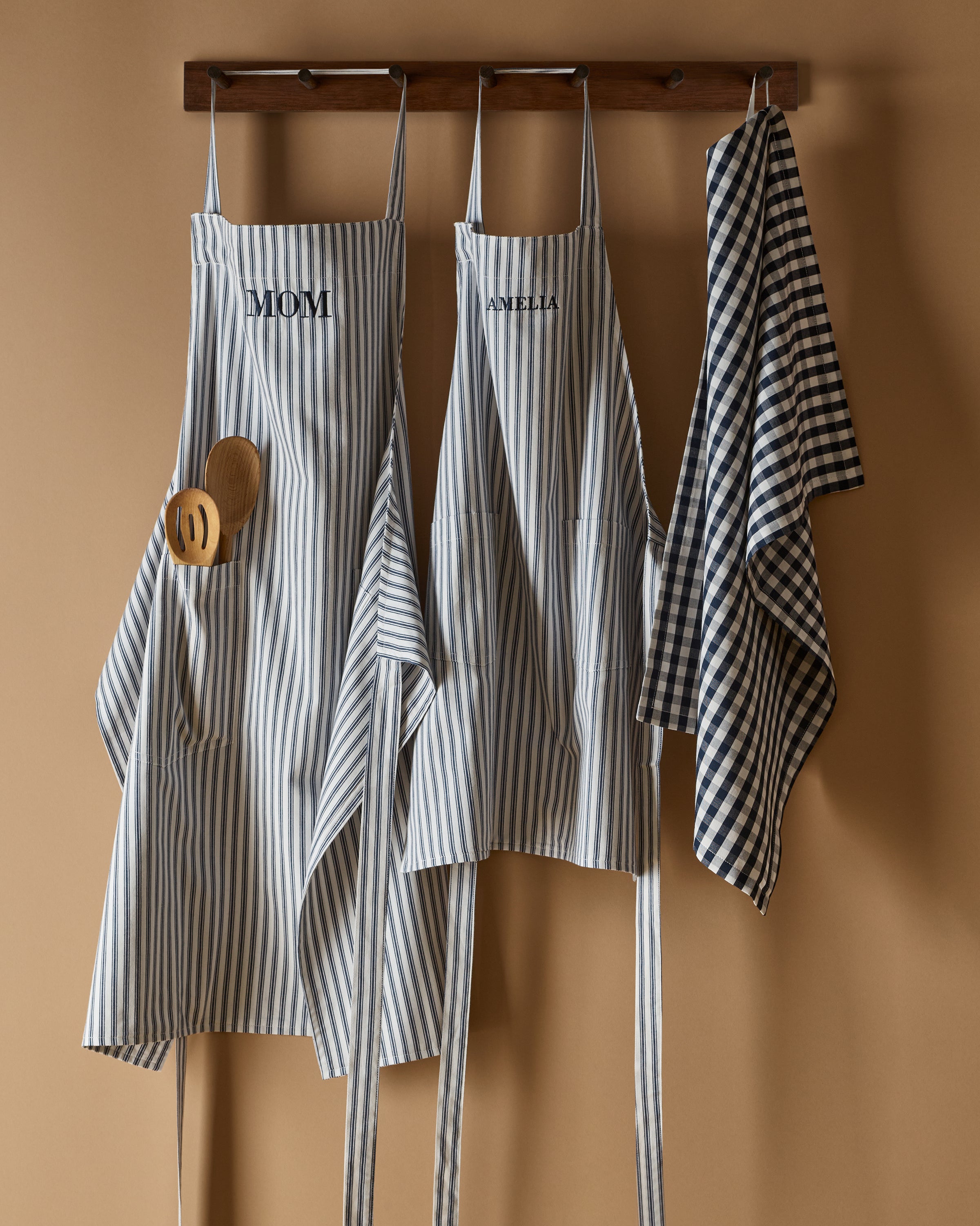 Three Petite Plume Navy French Ticking aprons hang on a wooden rack against a beige wall. Two embroidered with MOM and ANNELLA add a modern touch. Perfect for holiday baking, wooden utensils are tucked into one pocket, with a dishtowel hanging beside them.