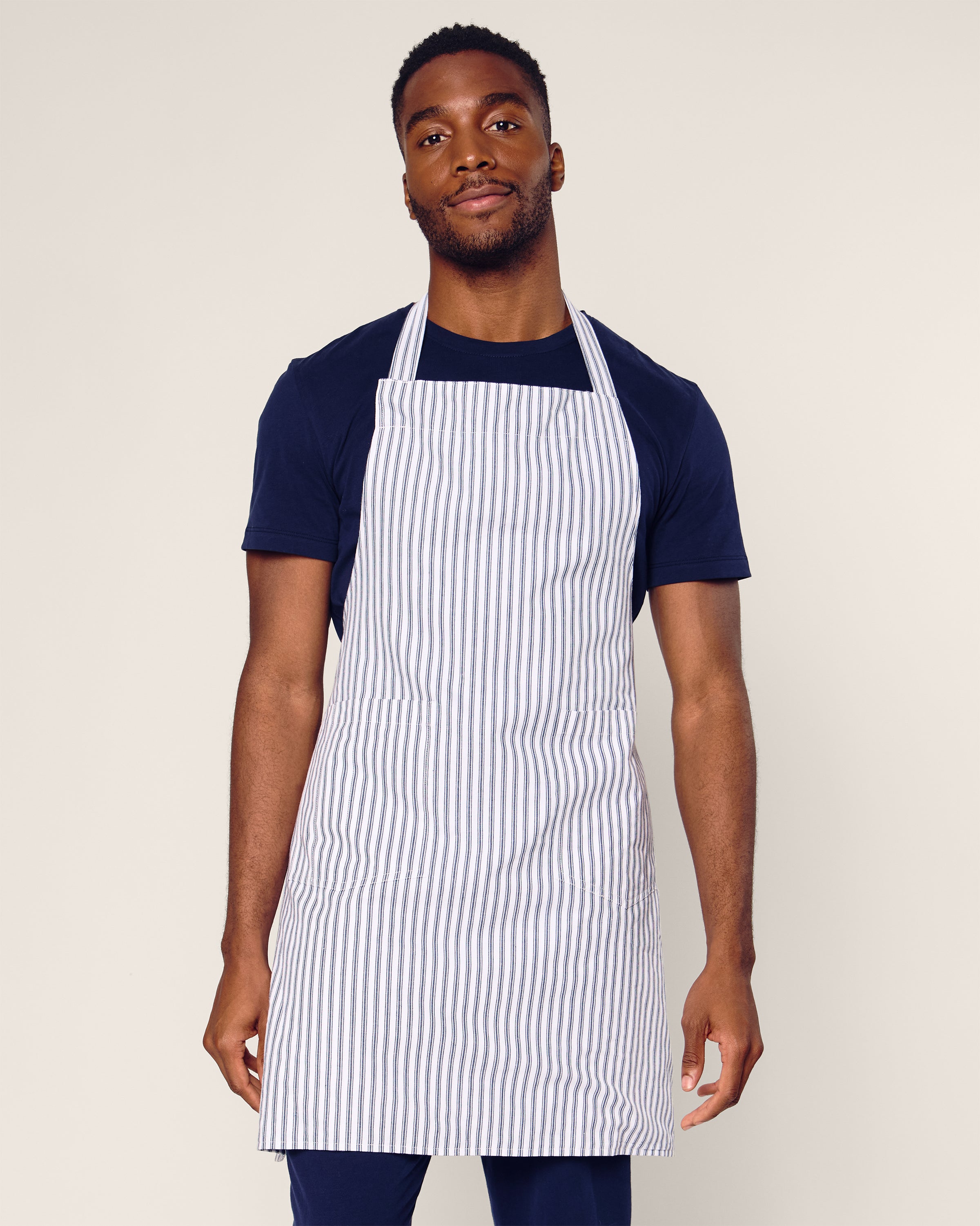 A person in a black t-shirt and Petite Plumes Adult Apron in Navy French Ticking stands against a plain light background, embodying modern holiday bakings essence while looking confidently at the camera.