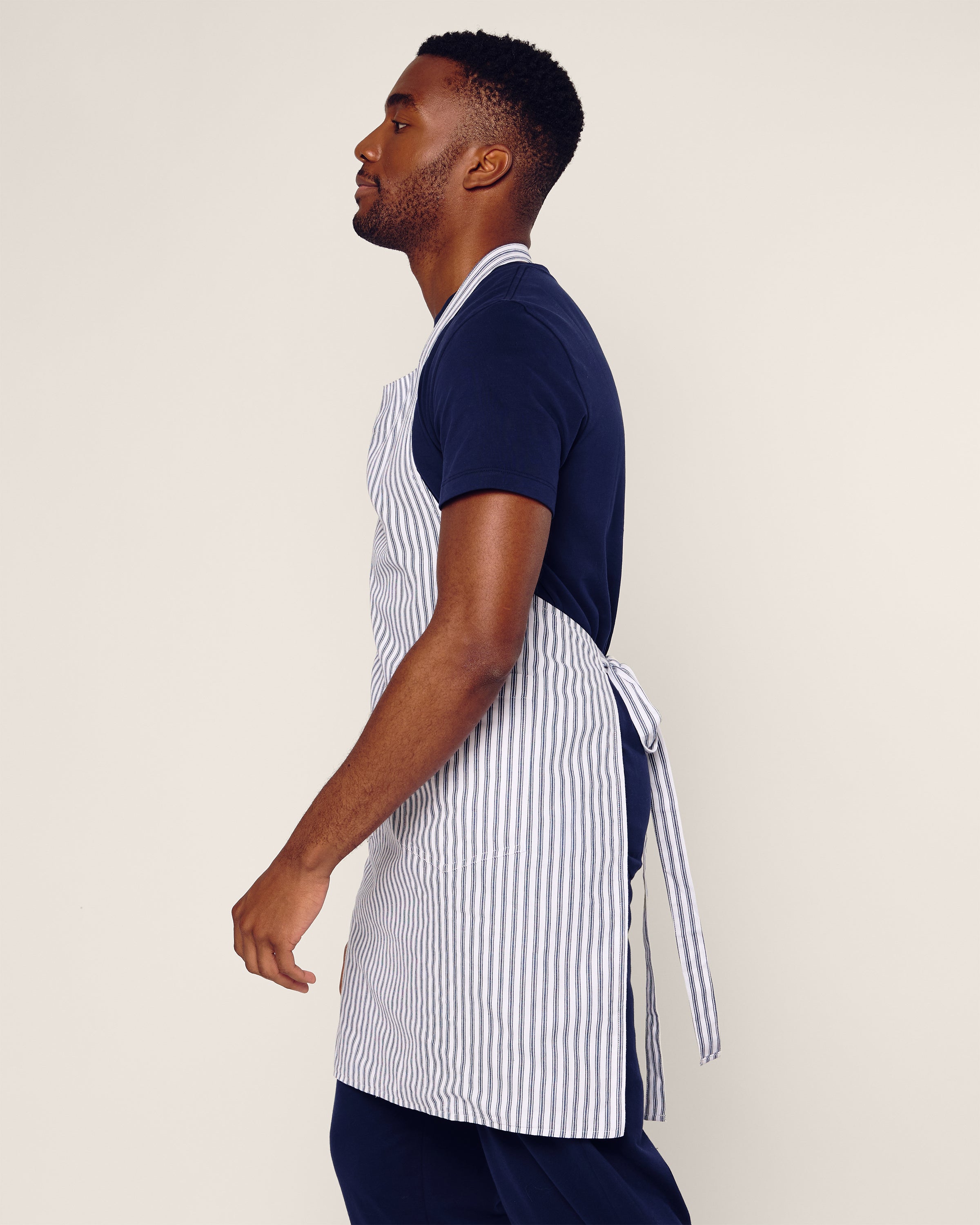 A man in a navy shirt wears the Petite Plume Adult Apron in Navy French Ticking, showcased from the side with its neatly tied back. The light background enhances the clean, modern vibe, ideal for holiday baking.