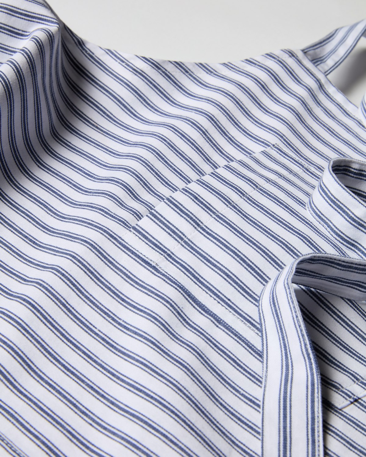 A close-up of the Petite Plume Adult Apron in Navy French Ticking reveals a modern fabric with blue and white vertical stripes, its slightly wrinkled surface offering a dynamic, textured appearance reminiscent of an apron ready for holiday baking adventures.