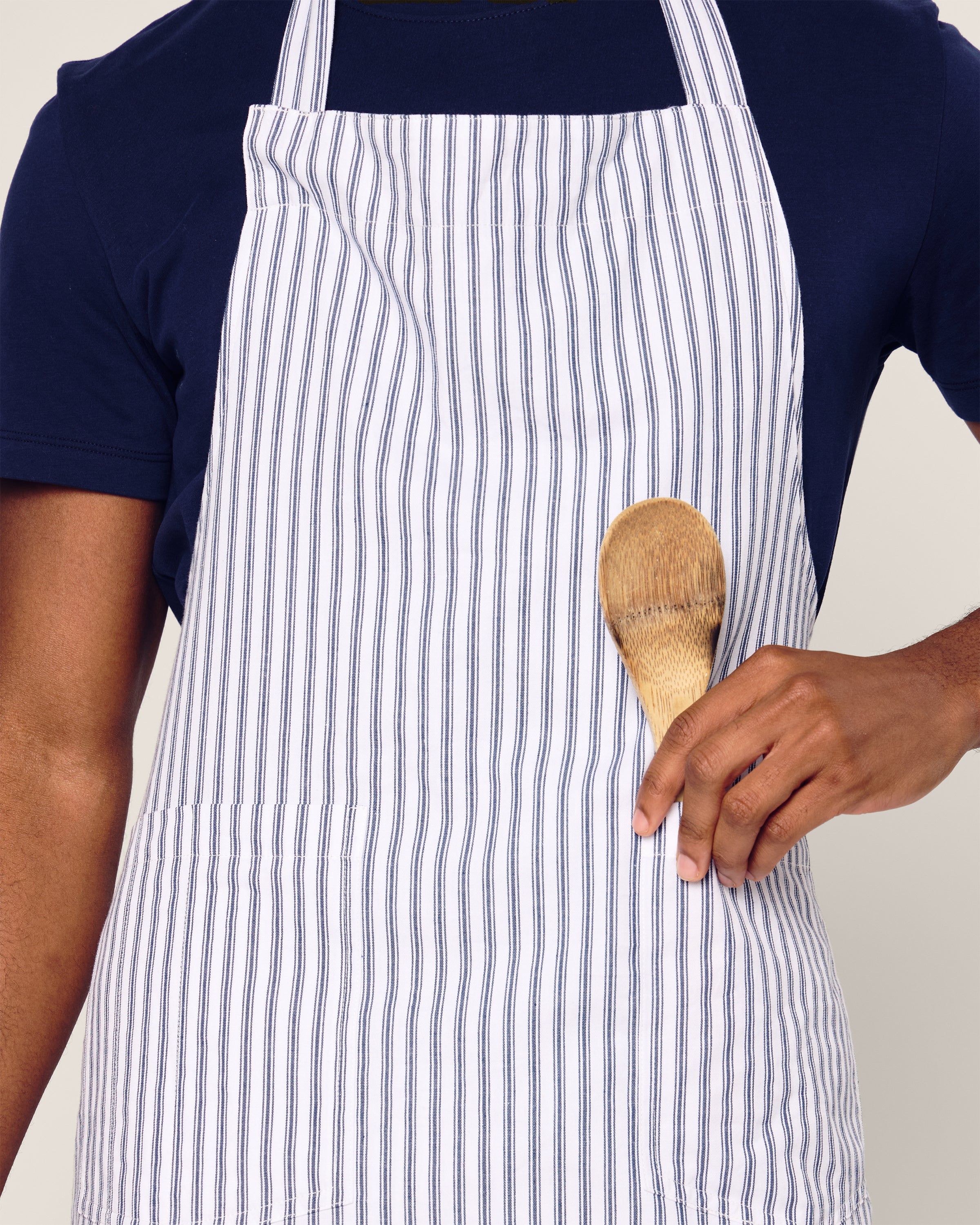 An individual engaged in holiday baking wears a modern blue shirt and the Petite Plume Adult Apron in Navy French Ticking, holding a wooden spoon against the apron. The image highlights their torso.