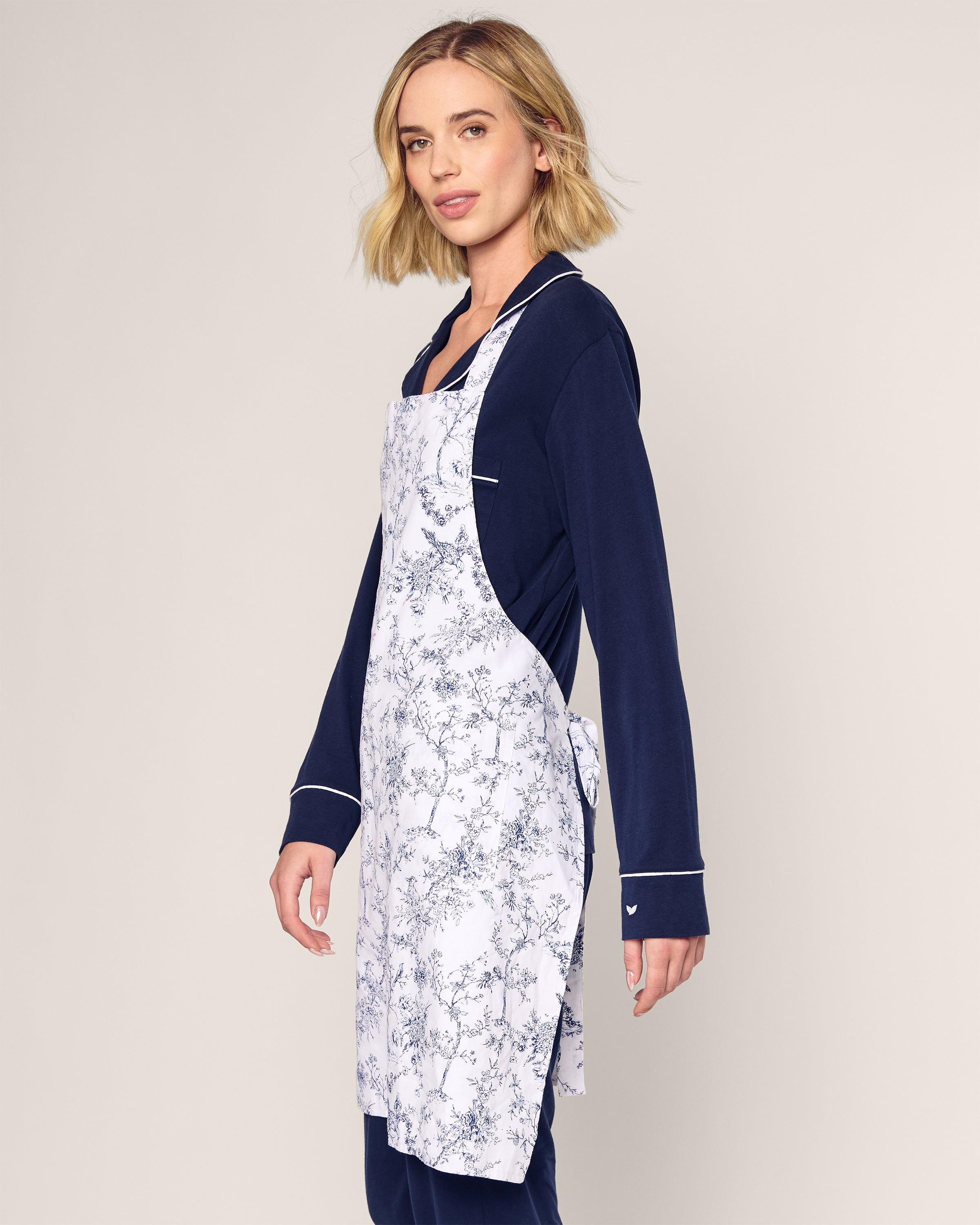 Wearing a long-sleeve navy outfit and Petite Plumes Adult Apron in Timeless Toile, featuring a white apron with a blue floral pattern, a person stands against a plain backdrop, exuding timeless beauty while looking slightly sideways.