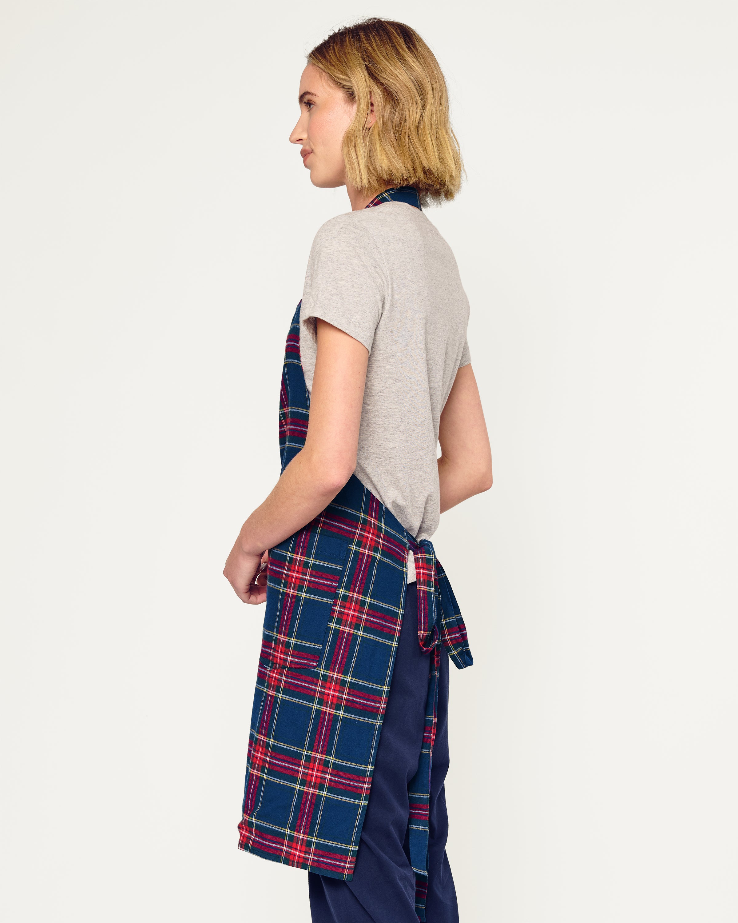 Two Adult Aprons in Windsor Tartan by Petite Plume, ideal for holiday baking, hang on a wooden rack beside a striped towel. One apron is labeled Dad, with a wooden spoon in its pocket. The background features a cozy warm beige wall.