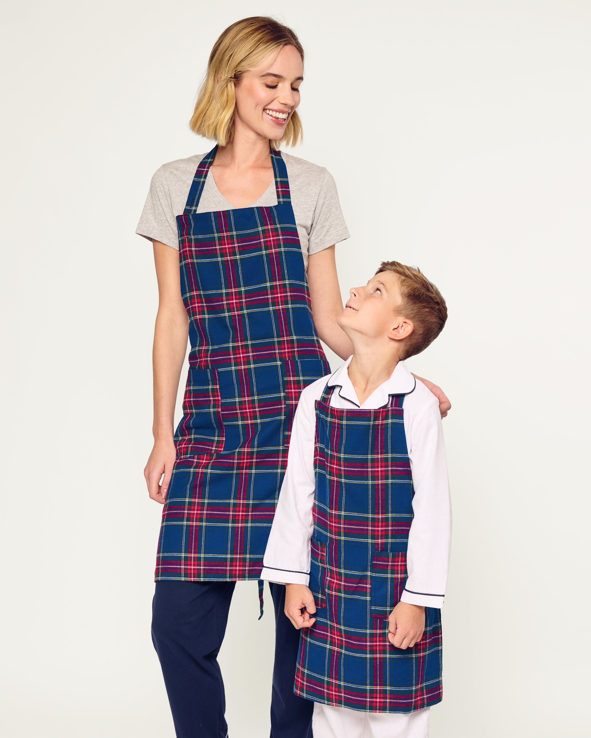 Two Adult Aprons in Windsor Tartan by Petite Plume, ideal for holiday baking, hang on a wooden rack beside a striped towel. One apron is labeled Dad, with a wooden spoon in its pocket. The background features a cozy warm beige wall.