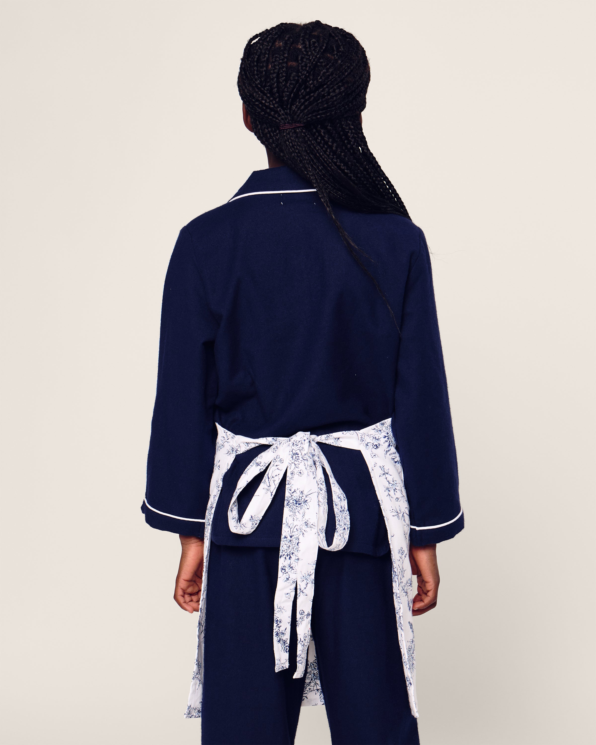A person with braided hair wears a dark blue long-sleeve top and matching pants, accented by Petite Plumes Kids Apron in Timeless Toile, tied around the waist. The backdrop is off-white.