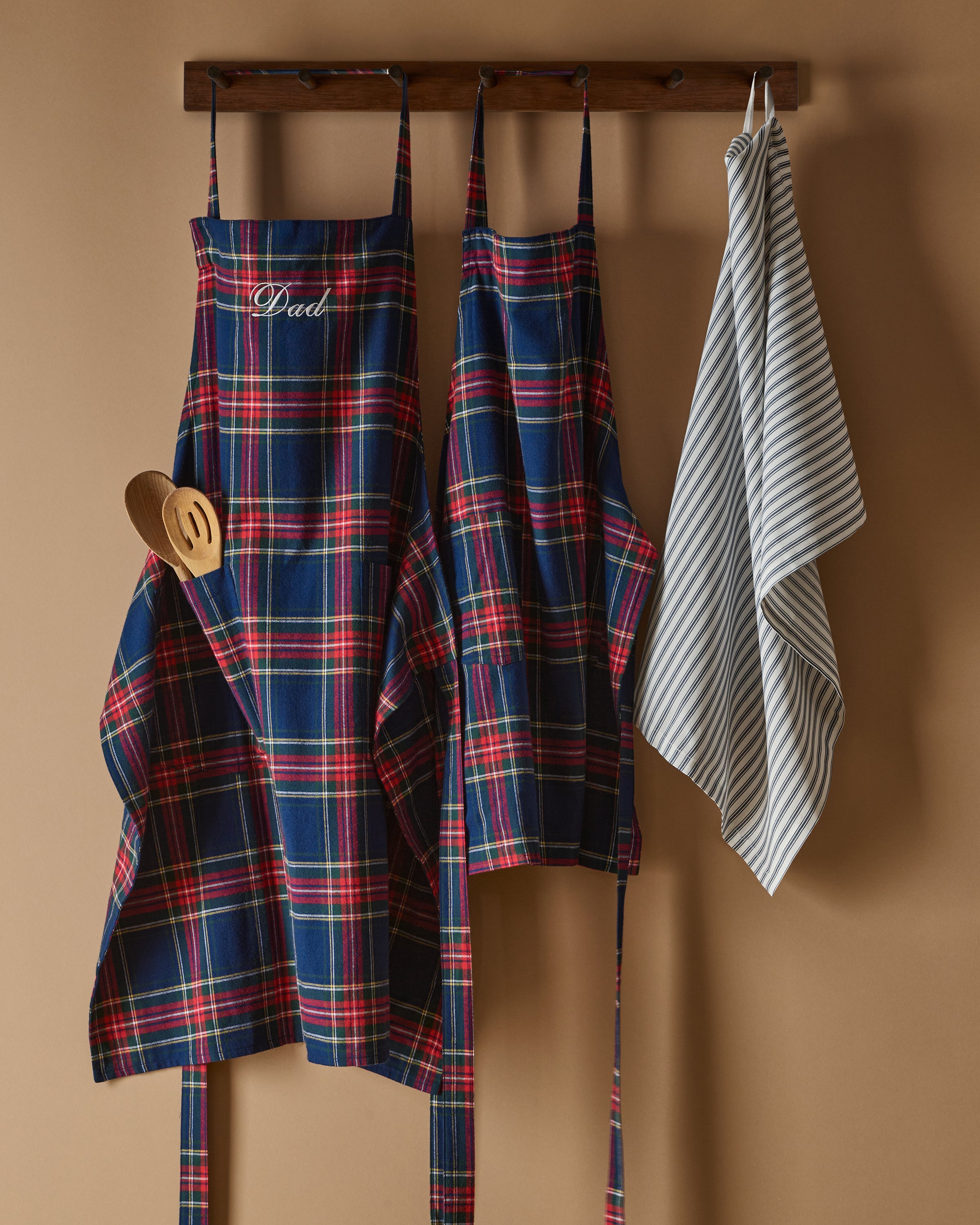 Two Windsor tartan kids aprons from Petite Plume hang on a wooden rack next to a striped dish towel, evoking holiday baking nostalgia. One apron, embroidered with Dad, holds a wooden spoon in its pocket against the warm beige wall backdrop.