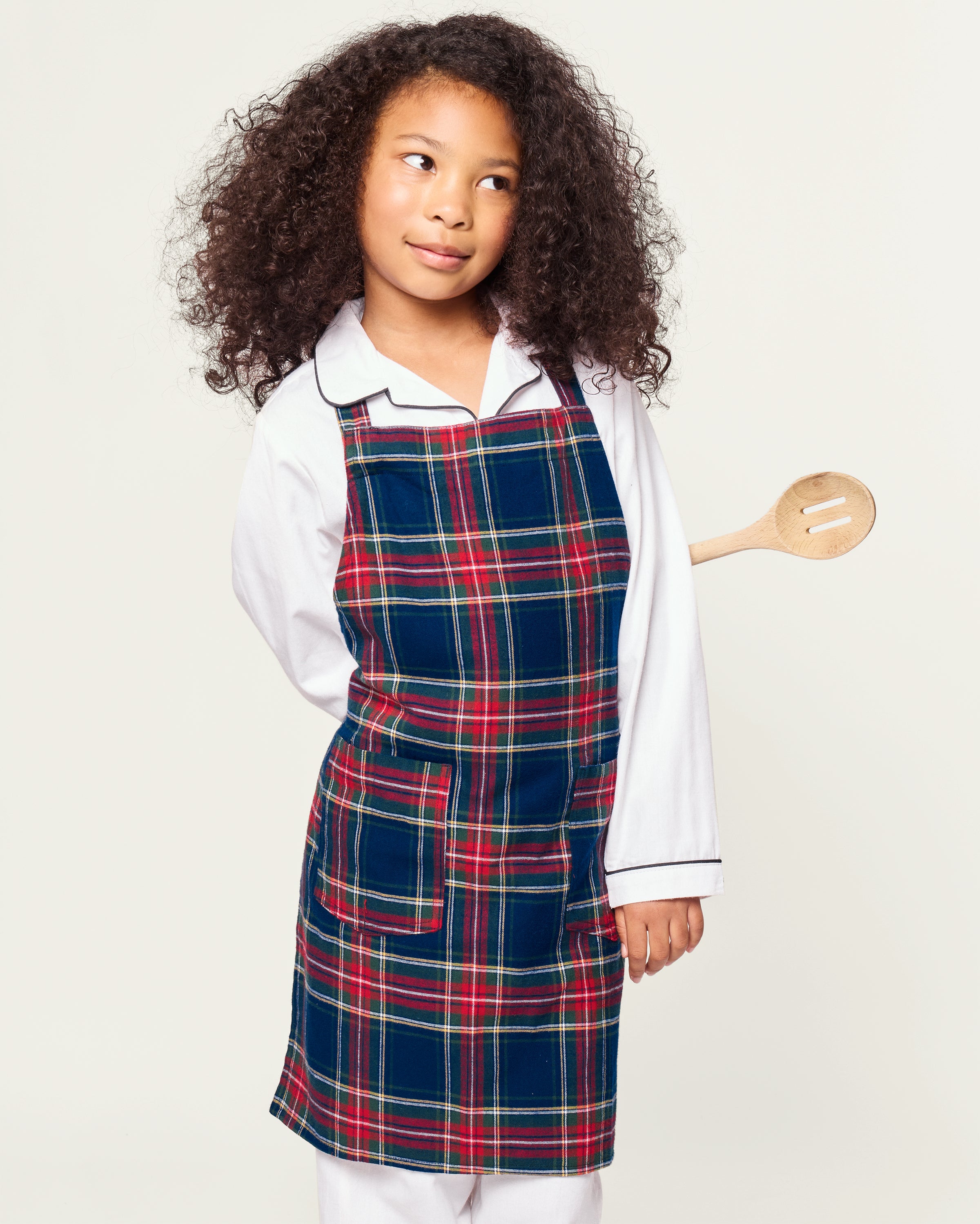 Two Windsor tartan kids aprons from Petite Plume hang on a wooden rack next to a striped dish towel, evoking holiday baking nostalgia. One apron, embroidered with Dad, holds a wooden spoon in its pocket against the warm beige wall backdrop.