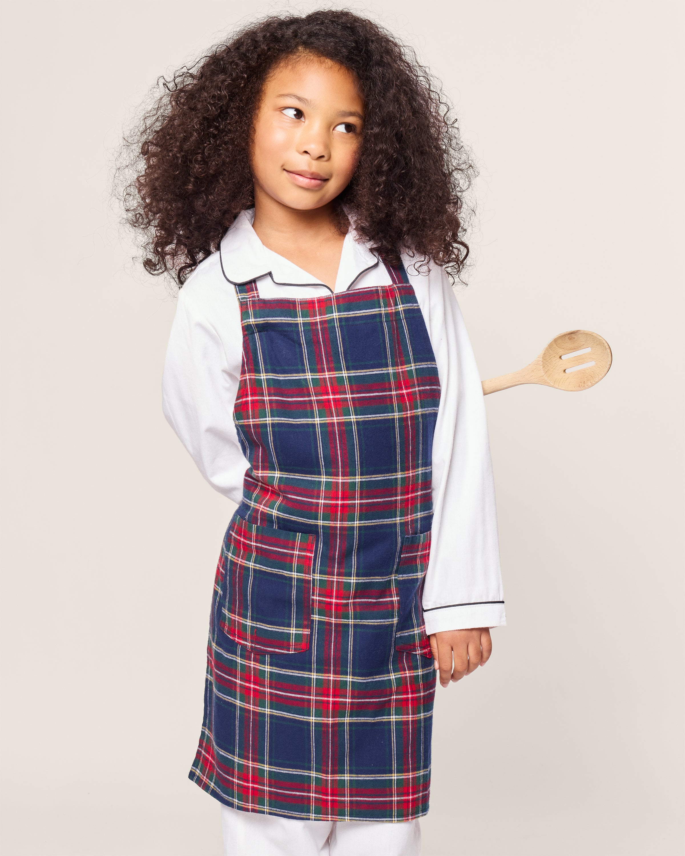 A curly-haired child, dressed in a white shirt and Petite Plumes Kids Apron in Windsor Tartan, playfully holds a wooden spoon behind their back, glancing to the side as if ready for holiday baking fun.