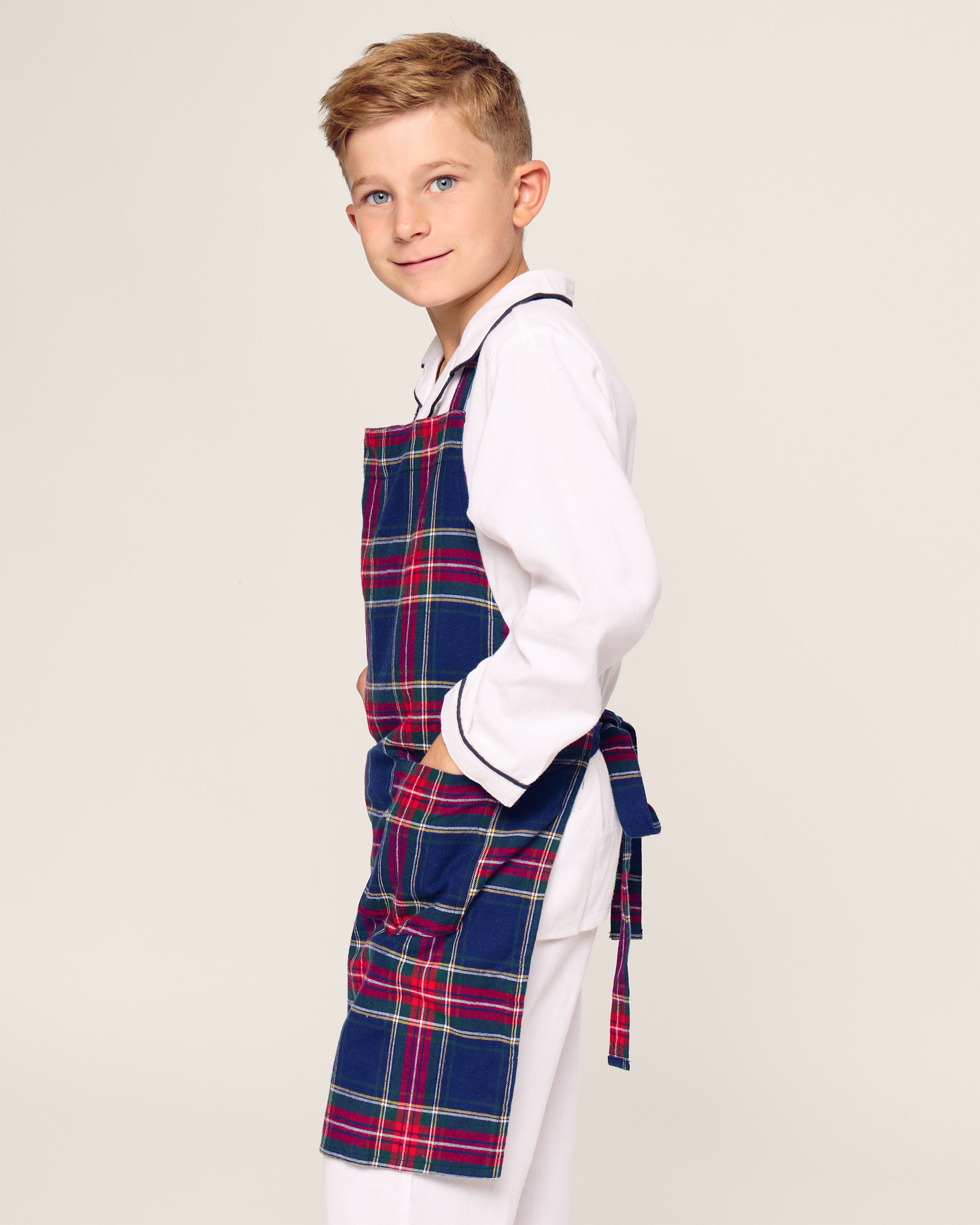 A young blond boy, wearing a white shirt and pants, models the Petite Plume Kids Apron in Windsor Tartan. He stands sideways with hands in the apron pockets, ready for holiday baking against a plain backdrop.