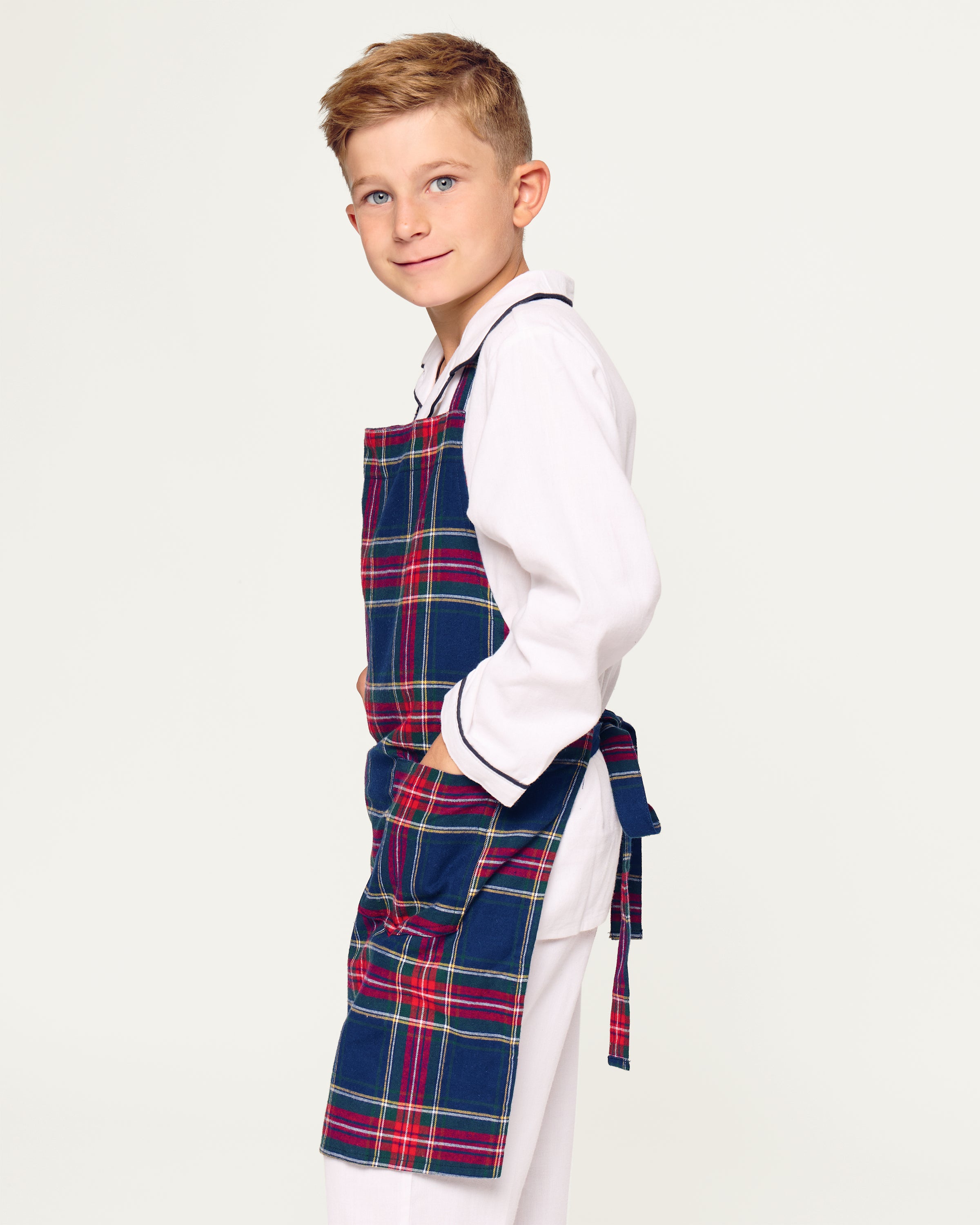 Two Windsor tartan kids aprons from Petite Plume hang on a wooden rack next to a striped dish towel, evoking holiday baking nostalgia. One apron, embroidered with Dad, holds a wooden spoon in its pocket against the warm beige wall backdrop.