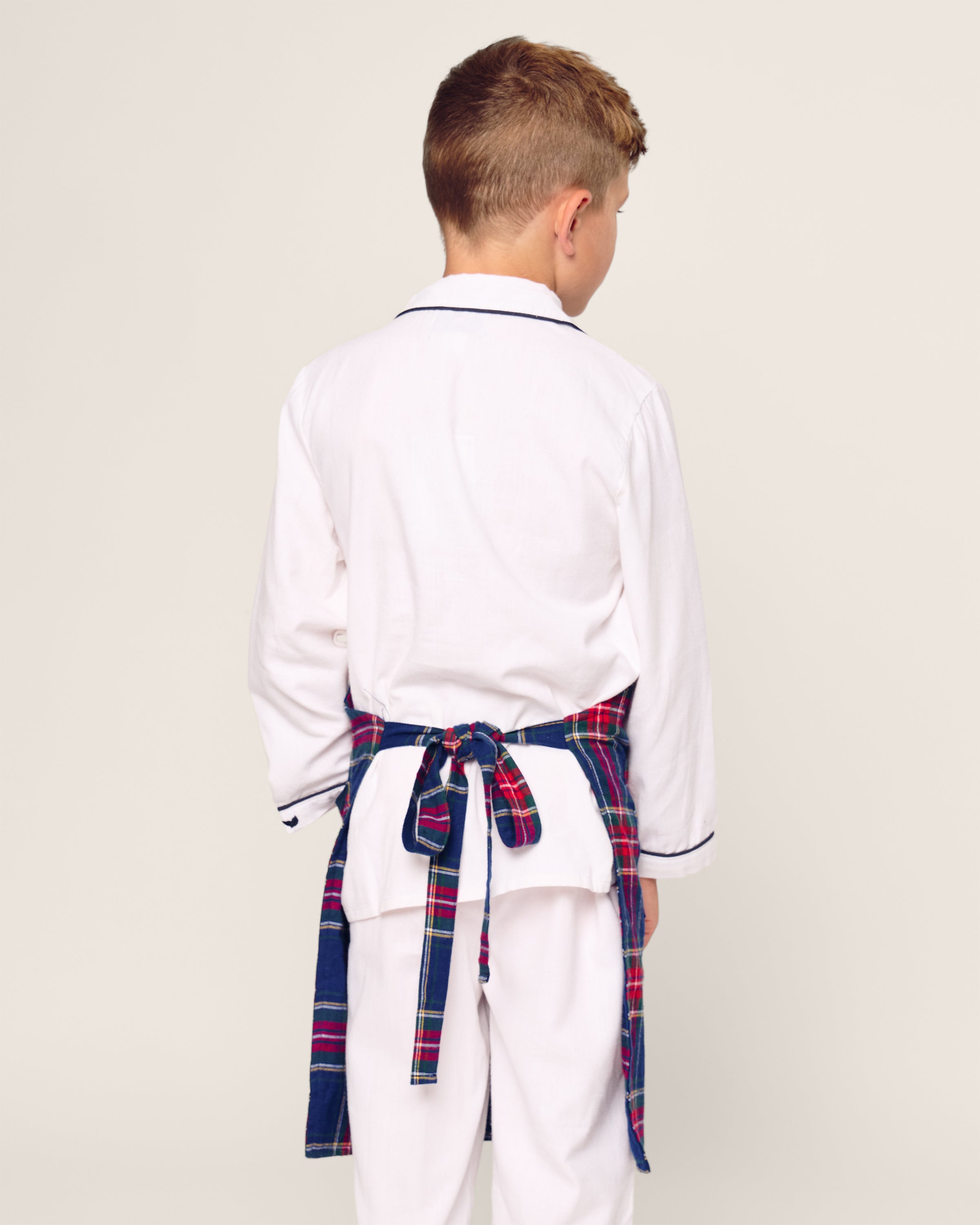 A young boy with short, light brown hair is wearing white pajamas with dark trim and Petite Plumes Kids Apron in Windsor Tartan. He faces away from the camera against a plain background, ready for holiday baking adventures.