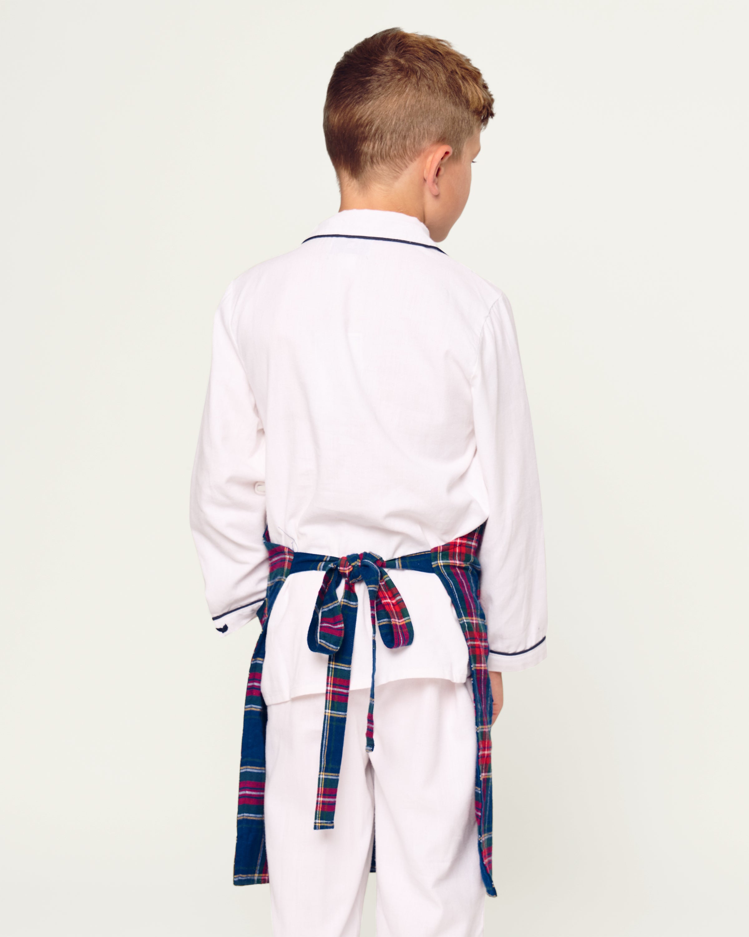 Two Windsor tartan kids aprons from Petite Plume hang on a wooden rack next to a striped dish towel, evoking holiday baking nostalgia. One apron, embroidered with Dad, holds a wooden spoon in its pocket against the warm beige wall backdrop.