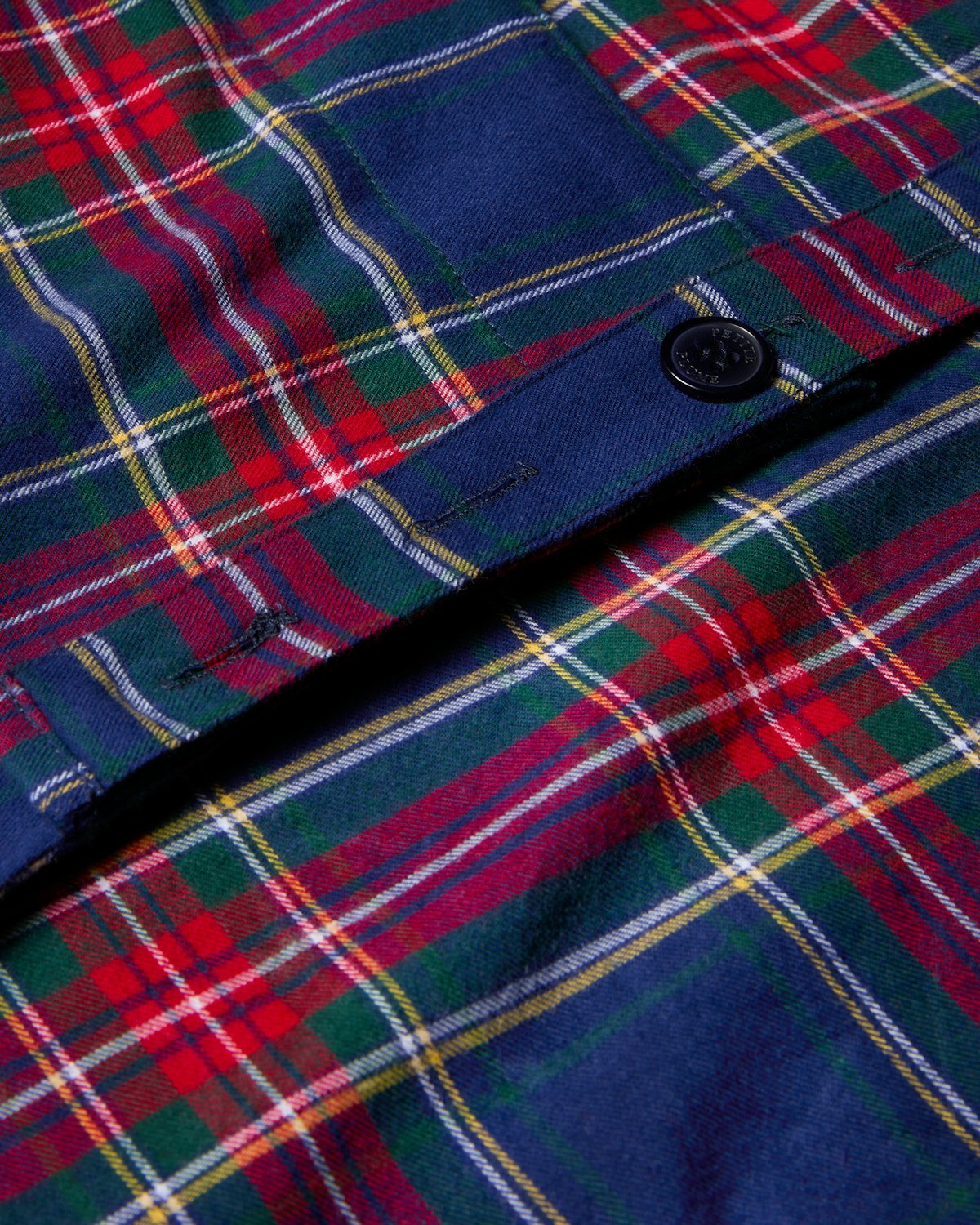 Close-up of the Kids Apron in Windsor Tartan by Petite Plume, featuring a festive plaid pattern of red, green, blue, and white stripes with a central black button—ideal for holiday baking.