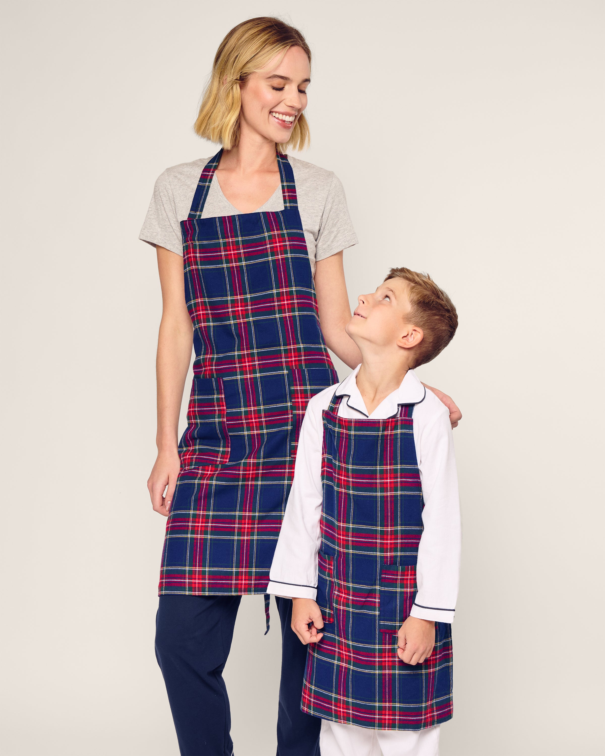 A woman and a boy share a joyful moment in matching Petite Plume plaid aprons, perfect for holiday baking. The woman smiles at the delighted boy. Their vibrant Kids Apron in Windsor Tartan stands out against the simple, light background.